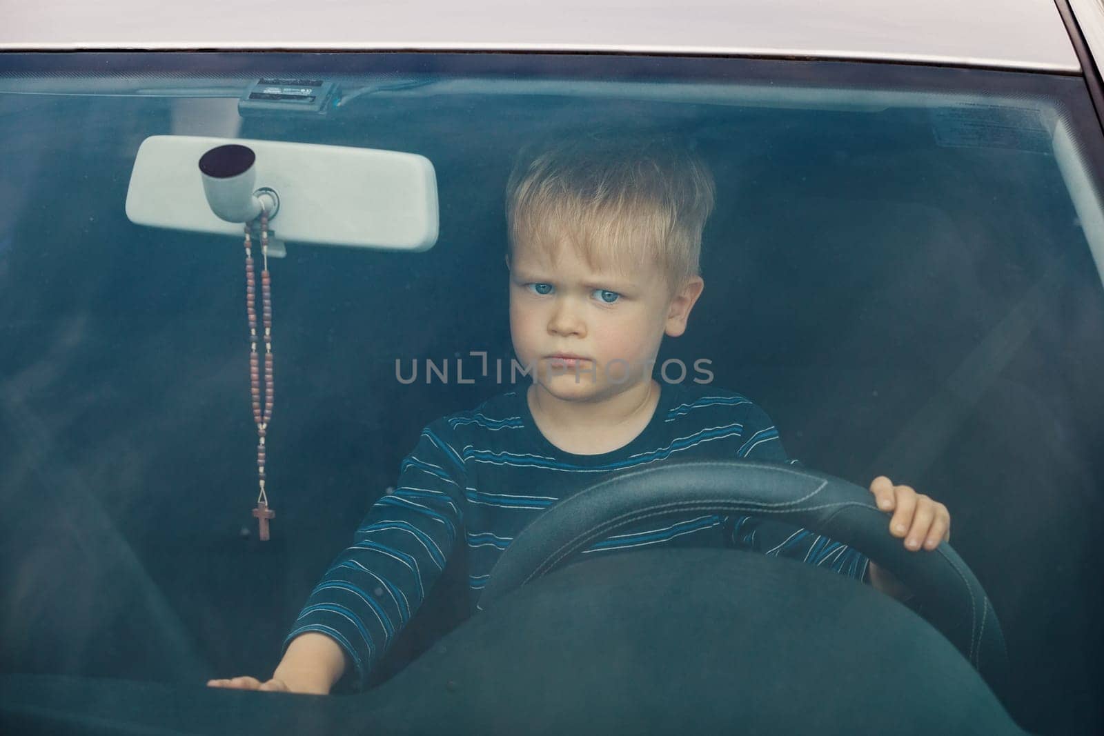 Portrait of a child sitting in a car behind the wheel from the front. The boy plays and imagines he is driving, he controls the radio with the other hand. by Lincikas