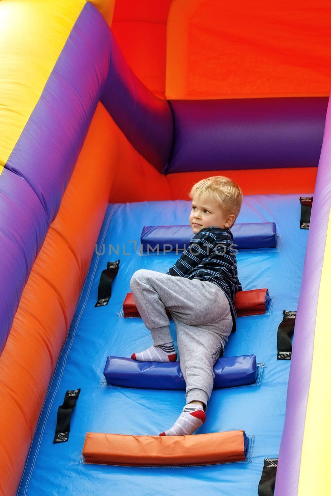 A cute boy climbs a colorful ladder on a high inflatable trampoline and looks into the distance.