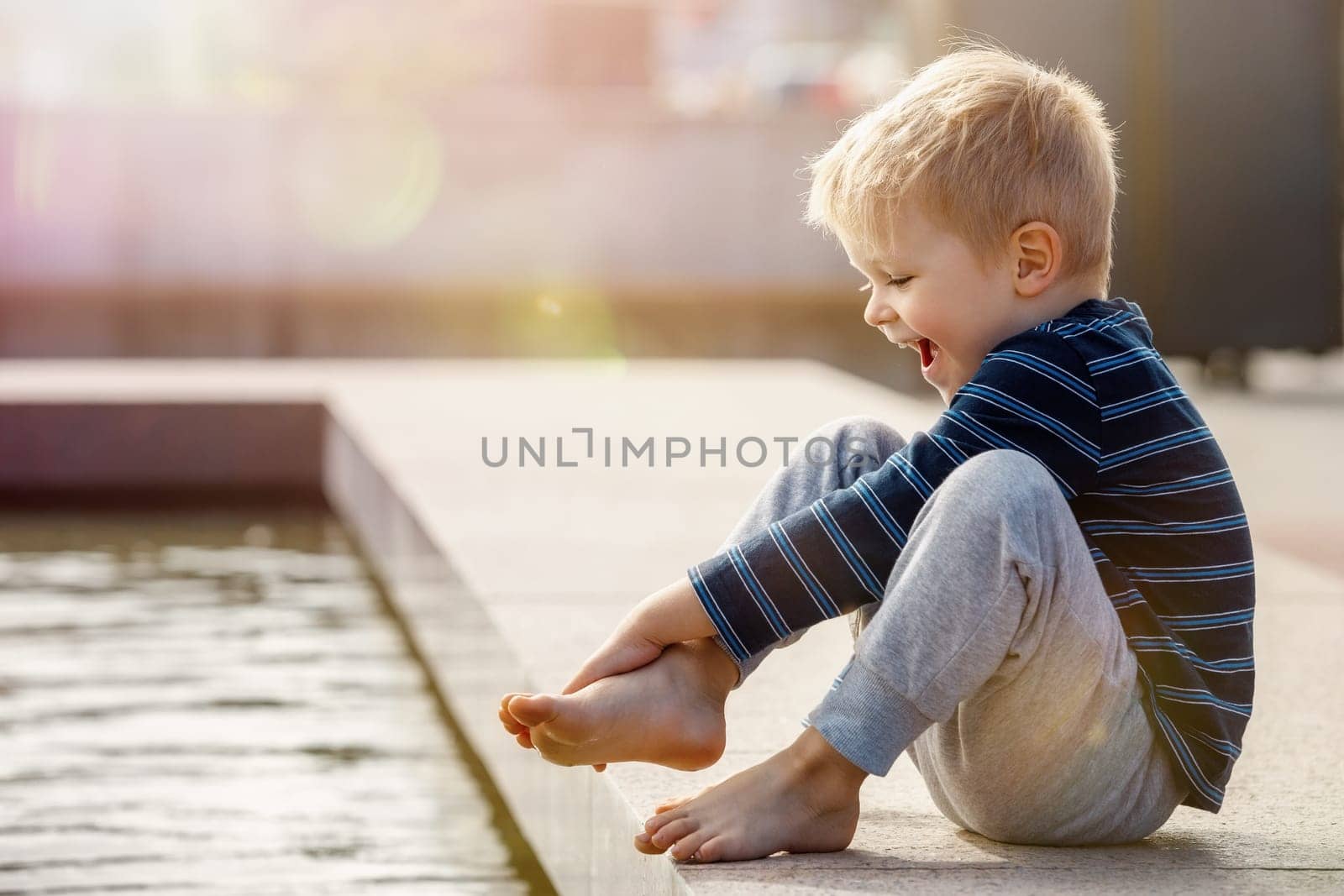 A curious child, a boy, barefoot touches the water of a city fountain. Hot and sunny summer day, the water is a good refreshment for the boy's feet. by Lincikas