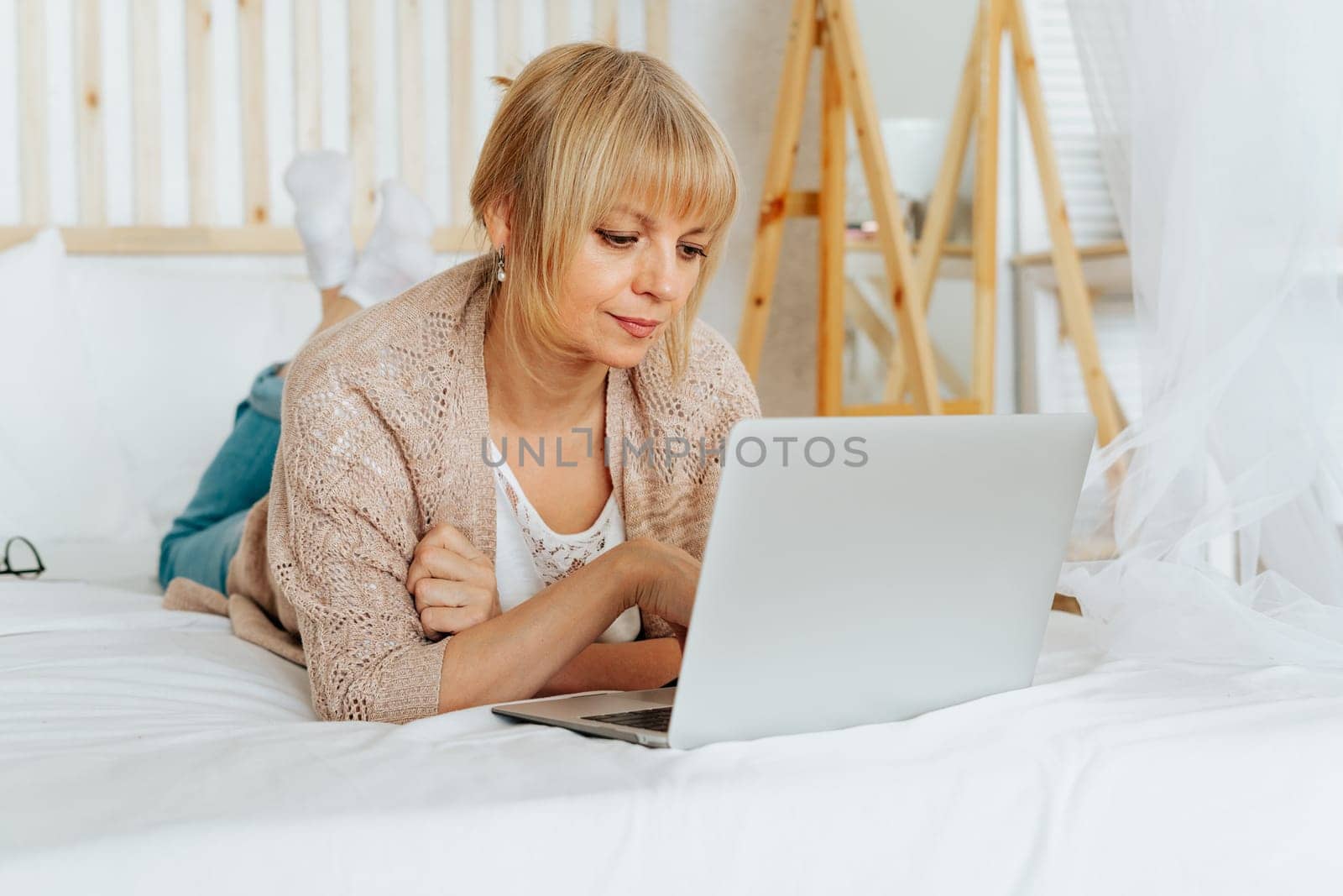 Senior mature woman lying on bed and surfing browse internet on her laptop. Adult female lady using computer for online shopping banking video conference call.