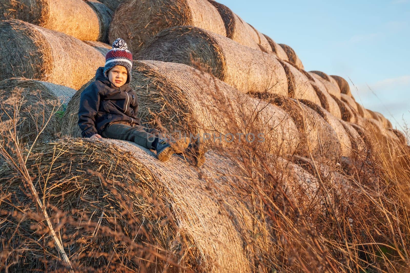A cute little boy in autumn clothes and a knitted hat poses against a background of golden hay bales. Horizontal photo.