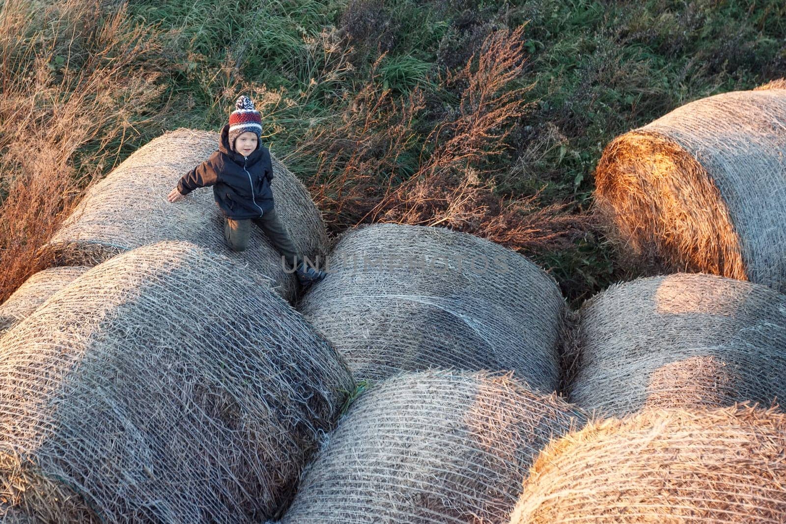 A little cute boy with a warm autumn hat among large rolls of straw in the evening sunlight, photo taken from above by Lincikas