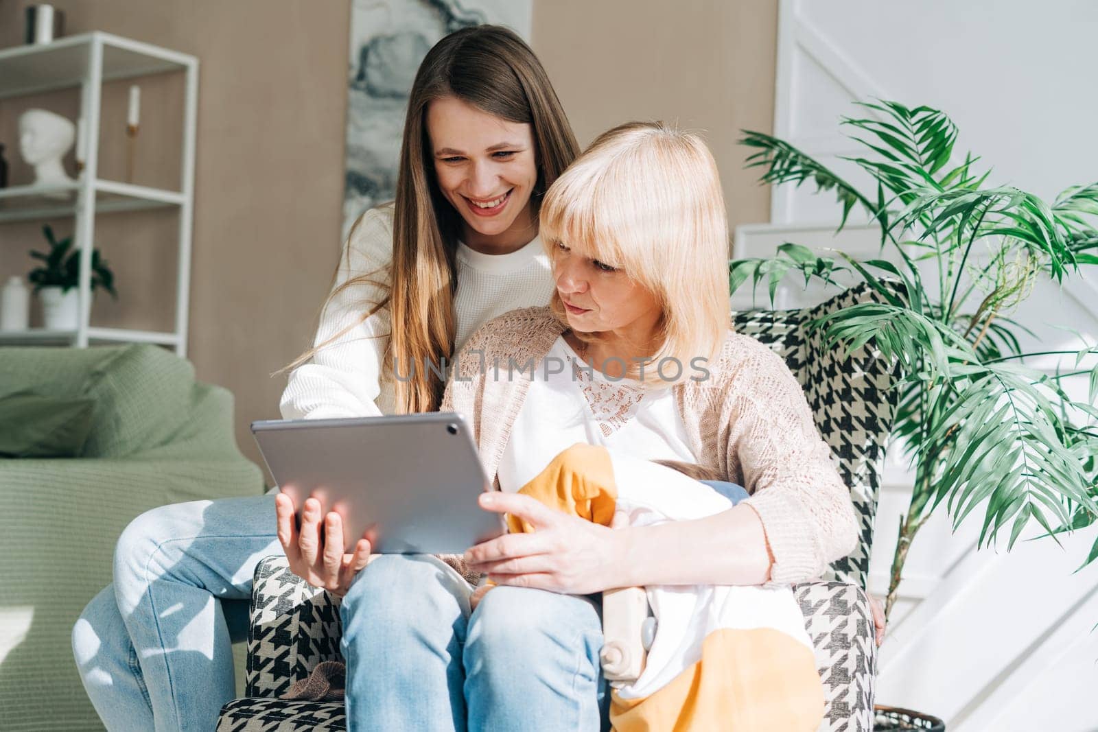 Senior woman and daughter playing with tablet pc. Smiling older woman and her granddaughter sitting home using gadget digital device for online shopping internet surfing video conference calling by Ostanina