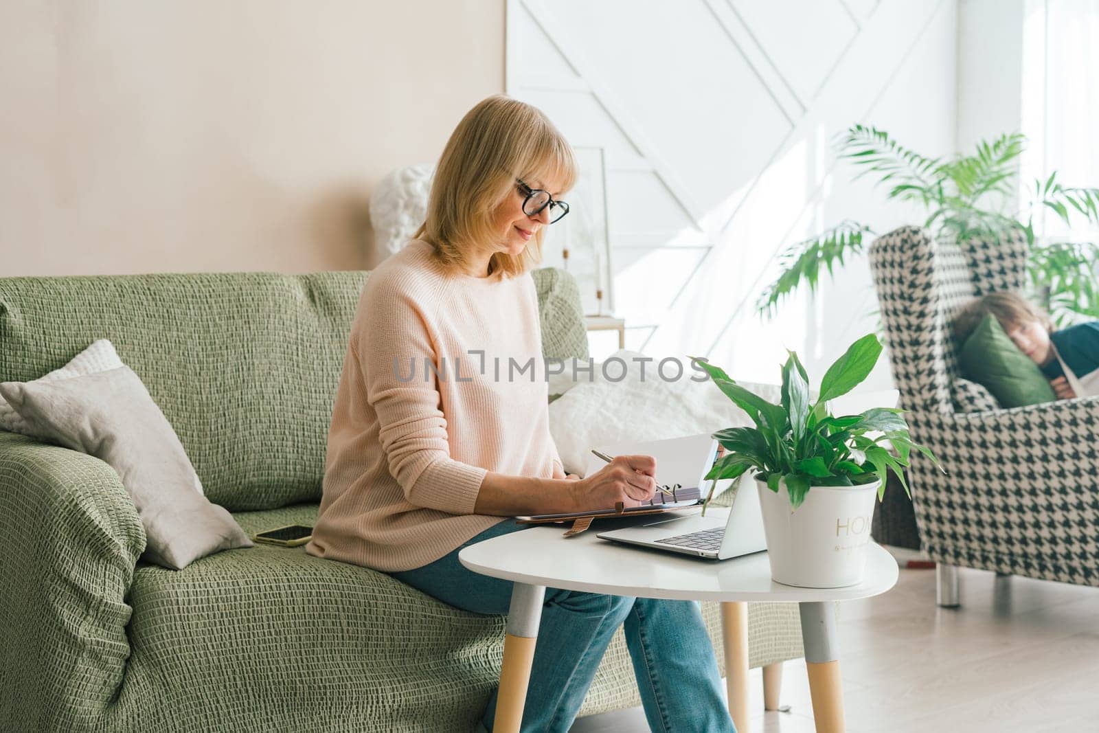 Adult mature woman lady writes a note in notebook notepad while doing online banking investments shopping on her laptop. Senior female uses laptop at home office. Copy space blank mockup by Ostanina
