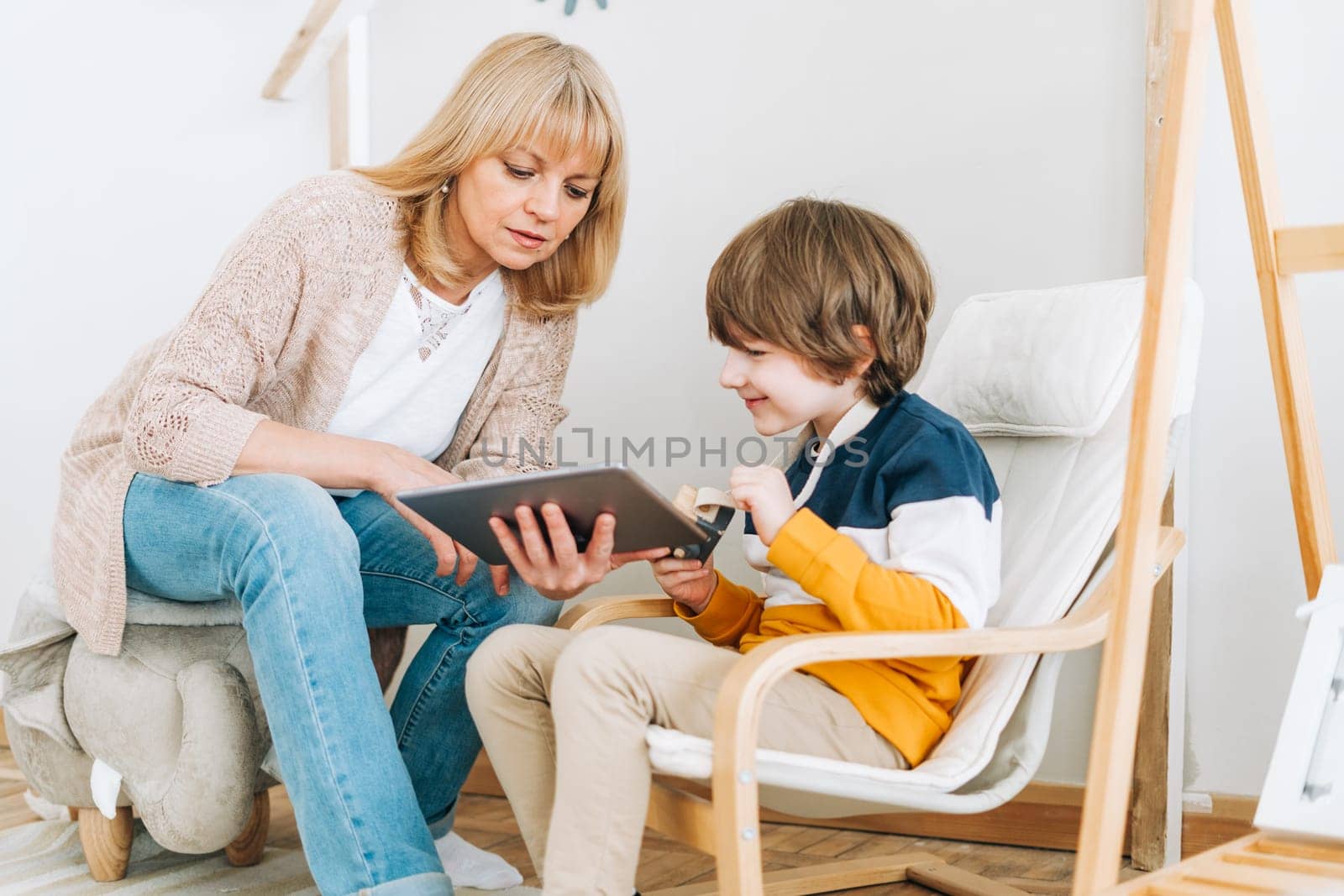 Happy caucasian grandmother sitting with grandson at home, looking in tablet computer device. Smiling family plays online video games with kid boy together. Watching on touch screen, relax.