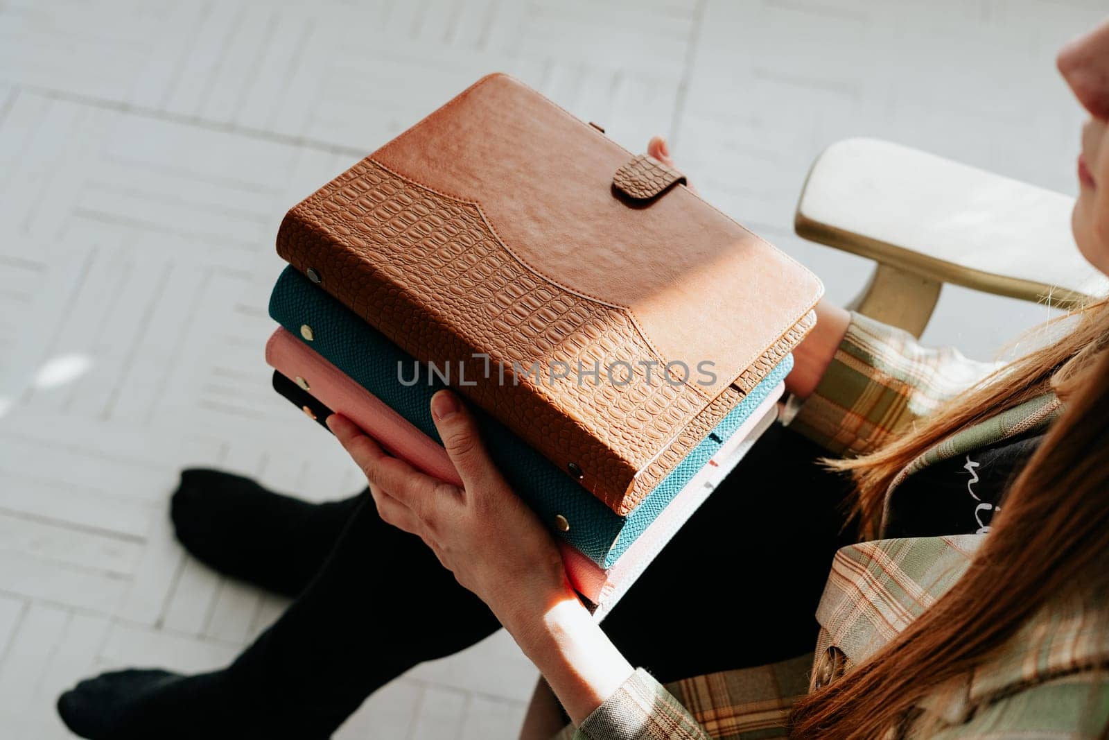 Cropped picture of young woman's hands holding stack of notebooks. Girl holds a pile of notepads books while sitting on a wooden chair. Blank space mockup books.