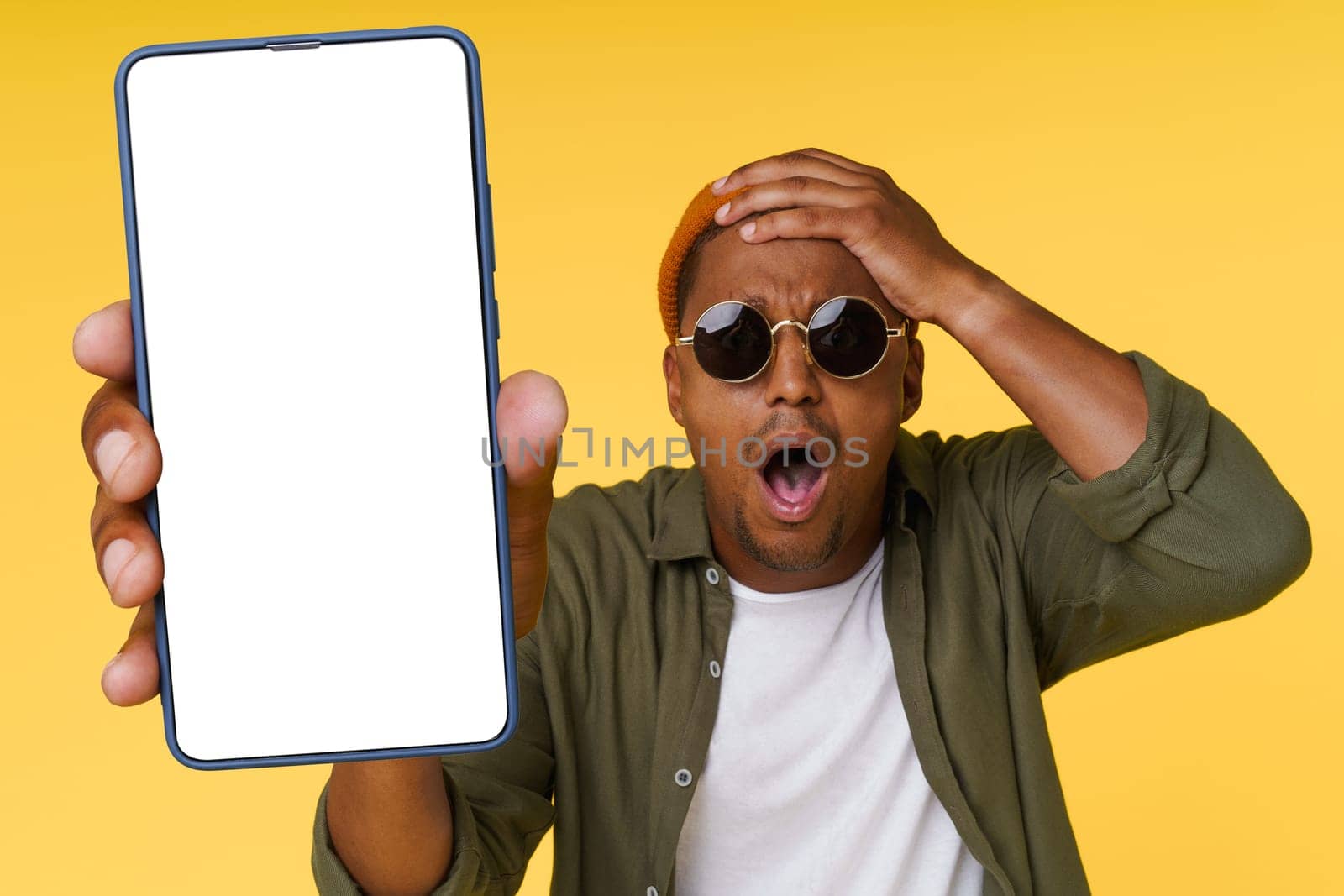 Surprised African man holds his head while showing phone with white blank screen on yellow background, concept for advertising. by LipikStockMedia
