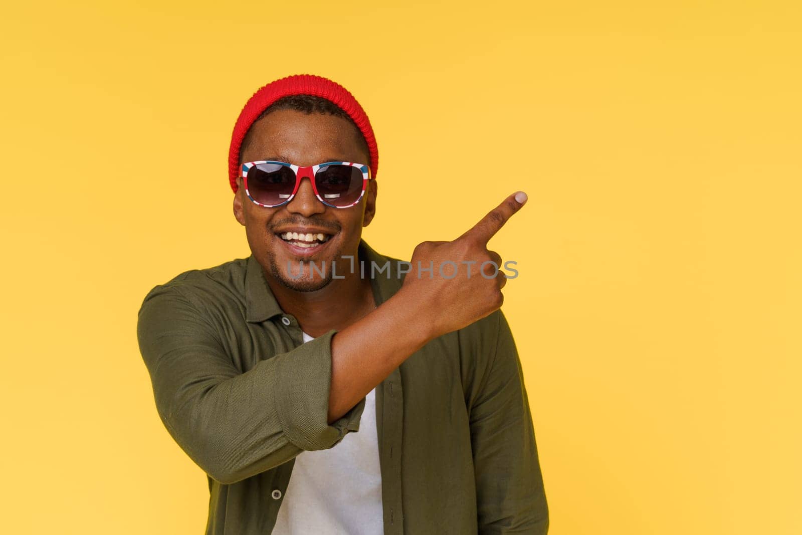 Smiling African man wearing sunglasses points to copy space on yellow background. Advertising concept. High quality photo