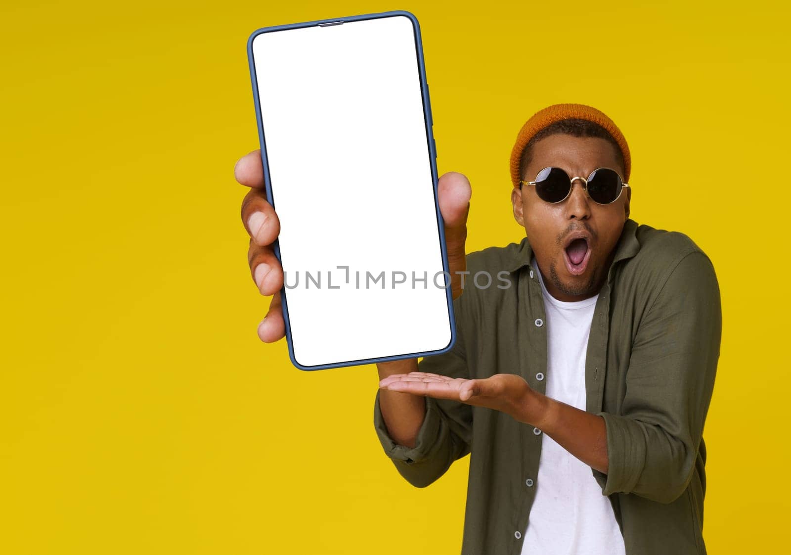 Surprised African man is holding phone with white blank screen against yellow background with copy space. The image is ideal for advertising concepts. High quality photo