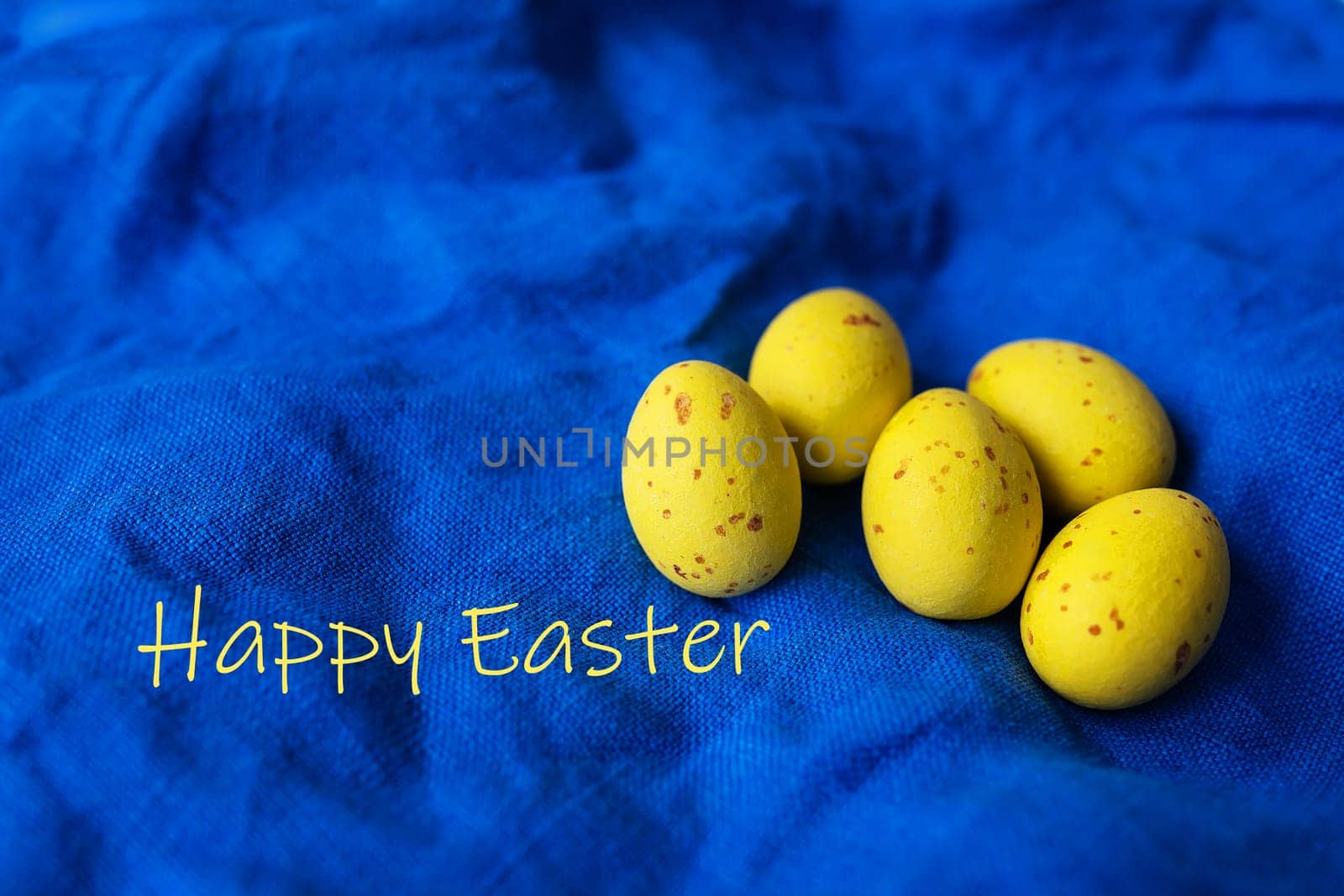 Top view of yellow decorated Easter eggs on napkin on blue textured linen background. Greeting card with the inscription Happy Easter