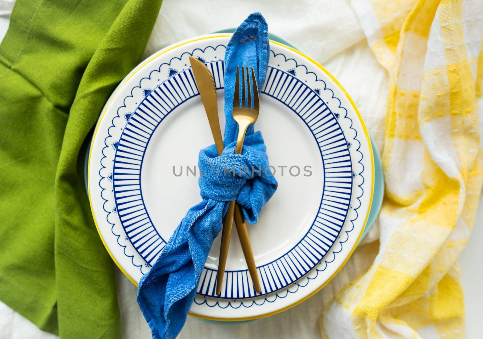 Gold cutlery is tied with a blue napkin, on which the plates are on a green-yellow linen napkin. Minimalist design, rustic. Birthday or wedding celebration. Serving design