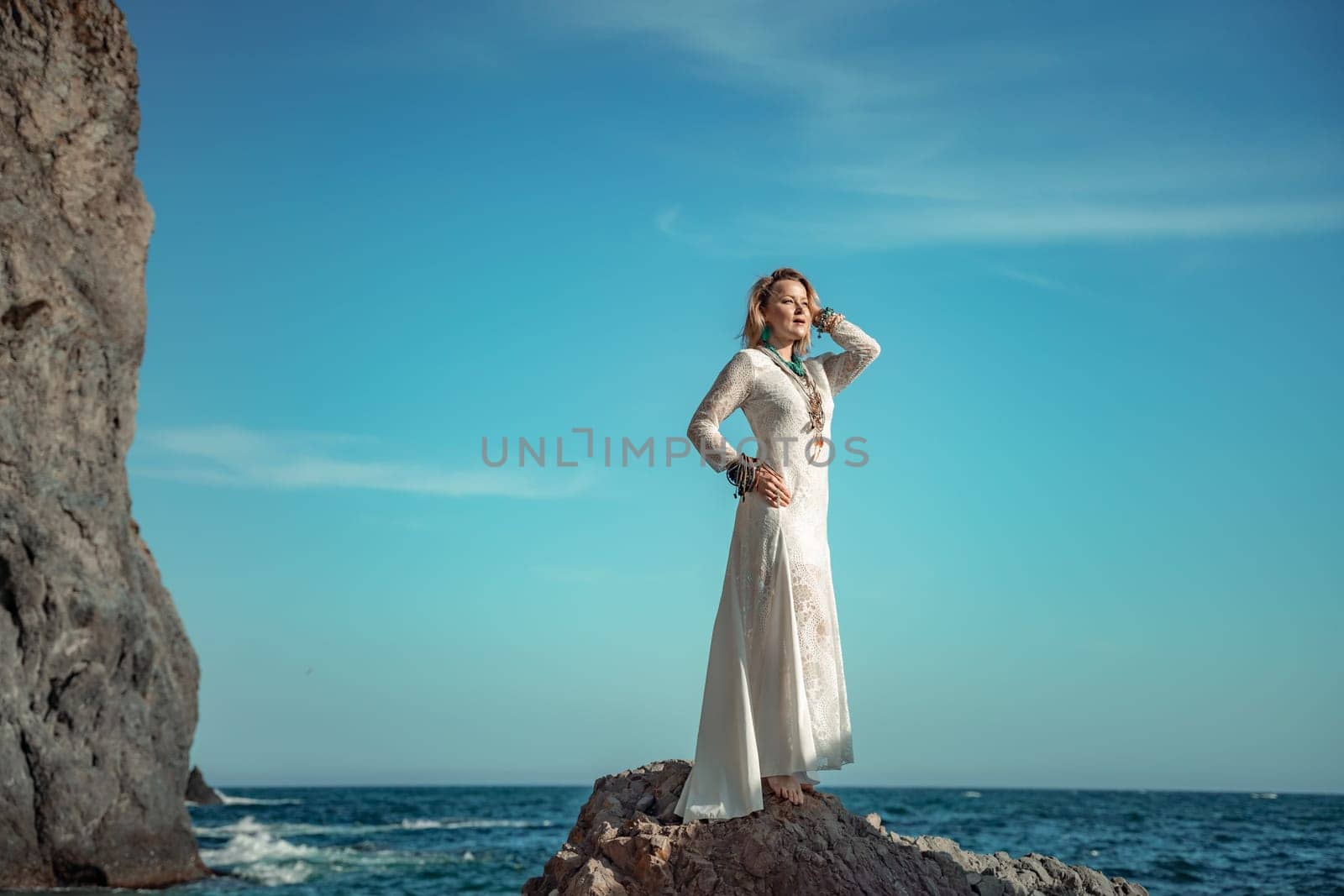 Woman white dress sea stones rocks.Middle-aged woman looks good with blond hair, boho style in a white long dress on beach jewelry around her neck and arms