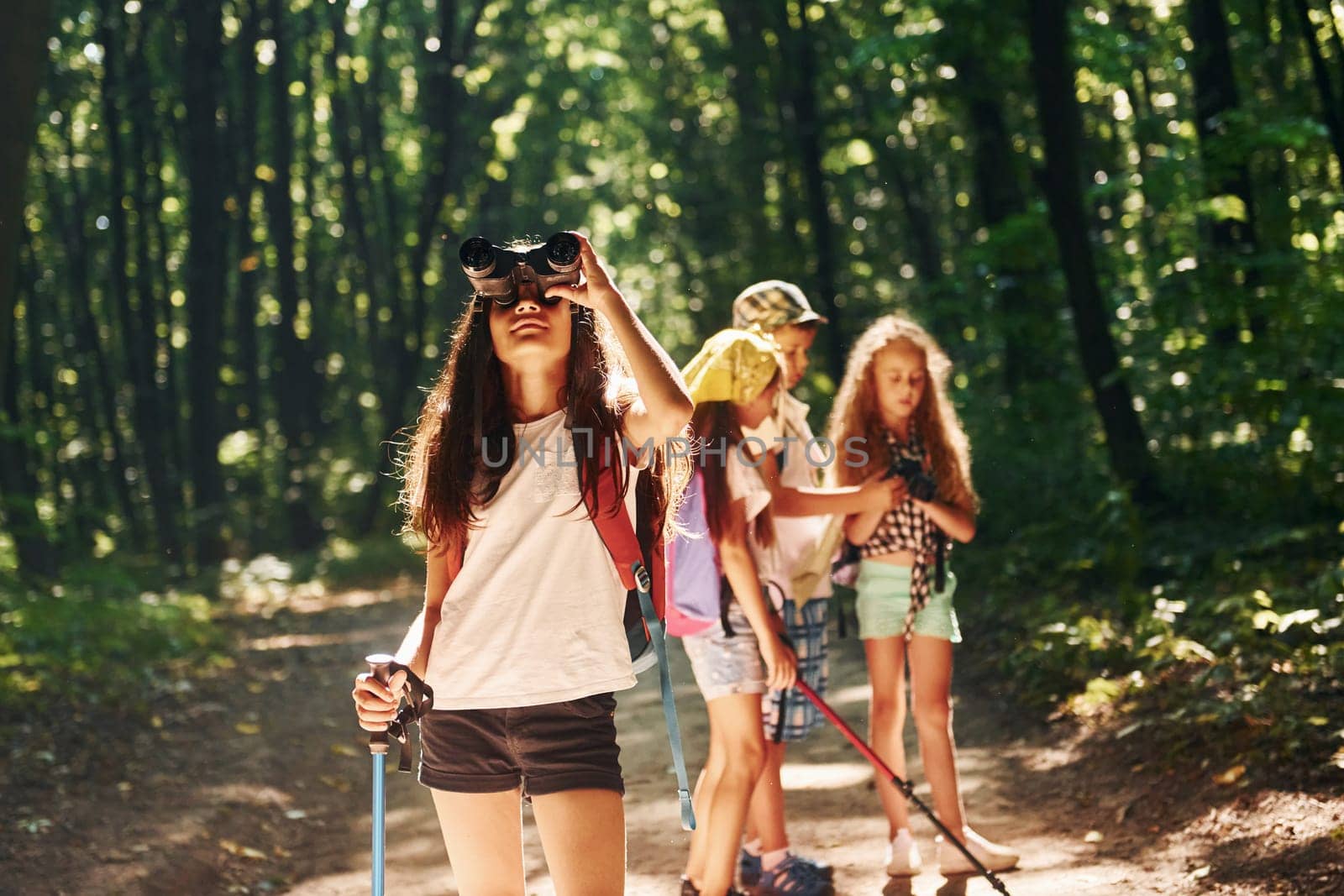 Girl standing in front of her friends. Kids strolling in the forest with travel equipment.