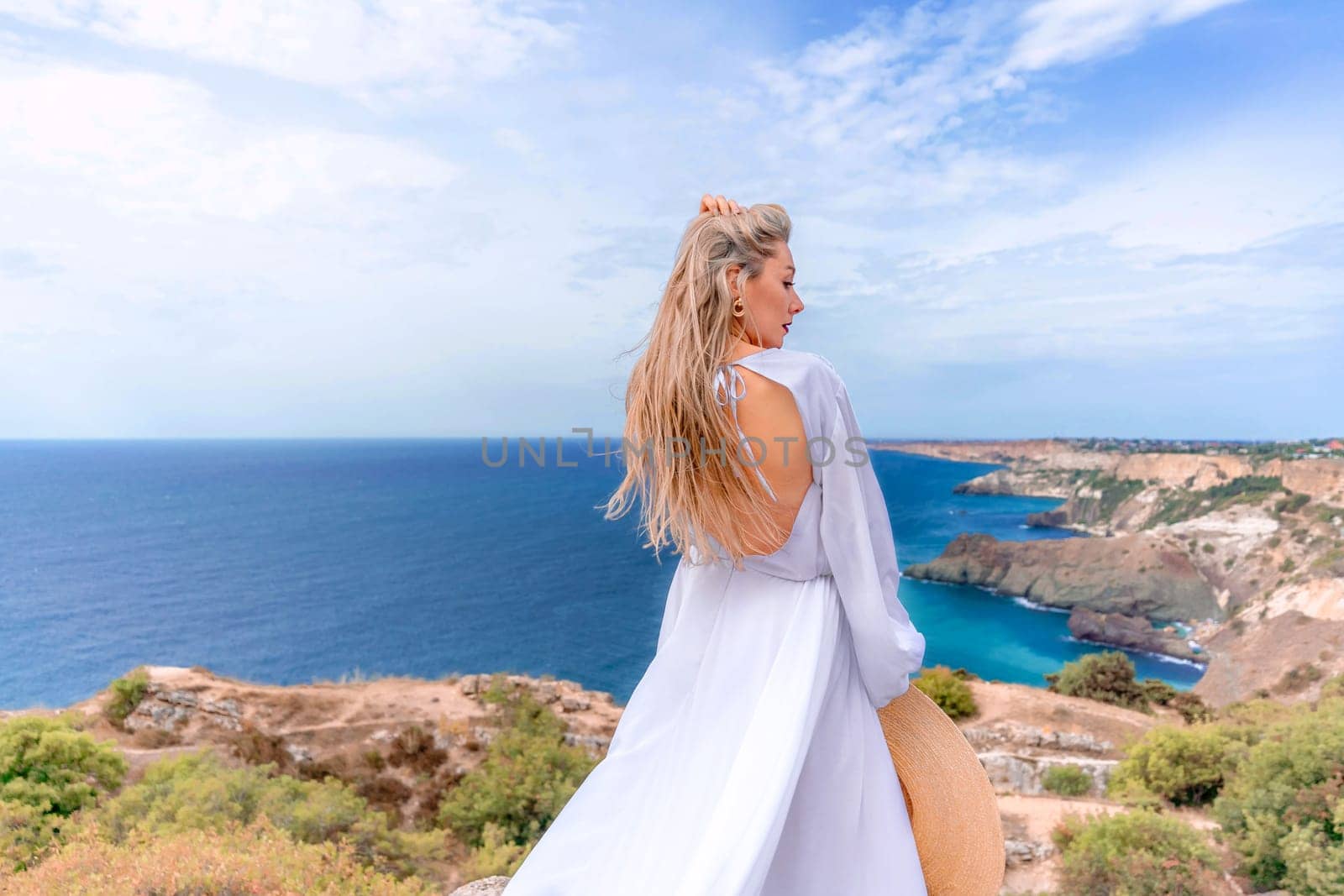 Blonde with long hair on a sunny seashore in a white flowing dress, rear view, silk fabric waving in the wind. Against the backdrop of the blue sky and mountains on the seashore