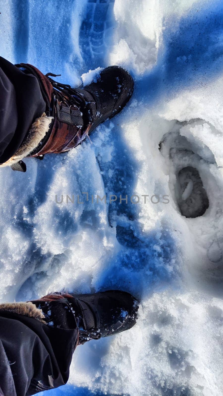 Feet of Hunter or fisherman in big warm boots on a winter day on snow. Top view. A fisherman on the ice of river, lake, reservoir on a spring day with melting ice. Dangerous fishing