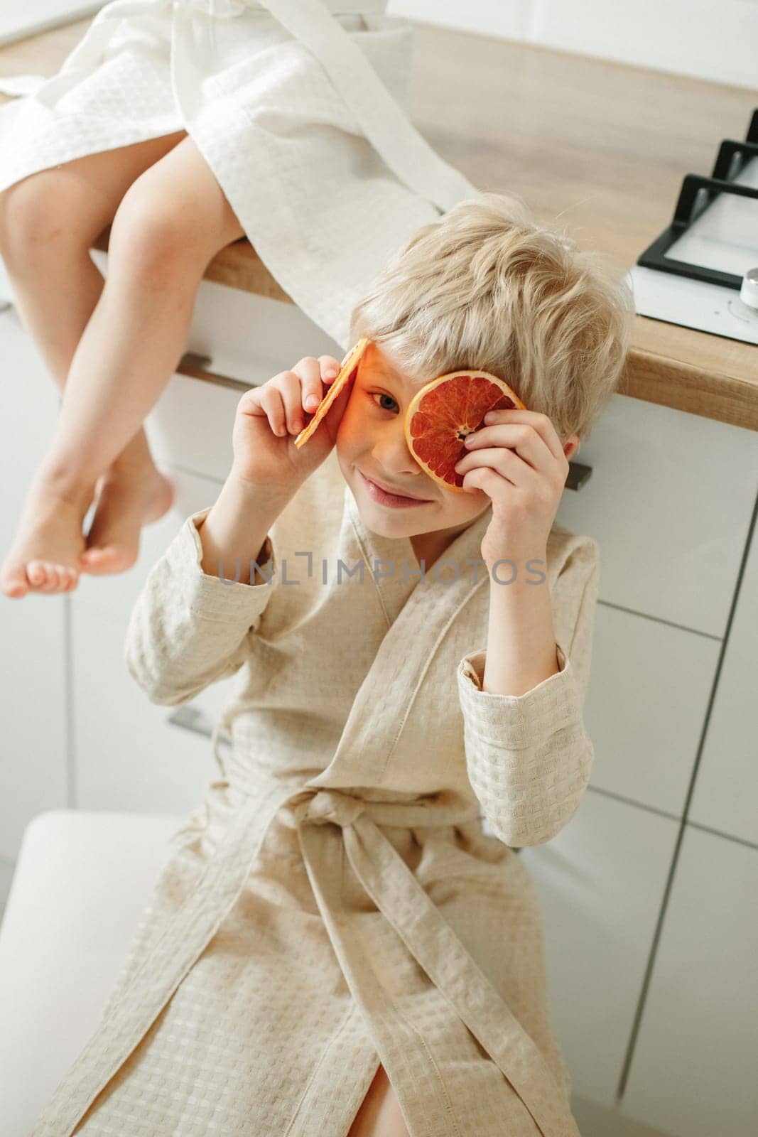 A boy in a bathrobe sits in the kitchen and holds candied oranges in his hands.