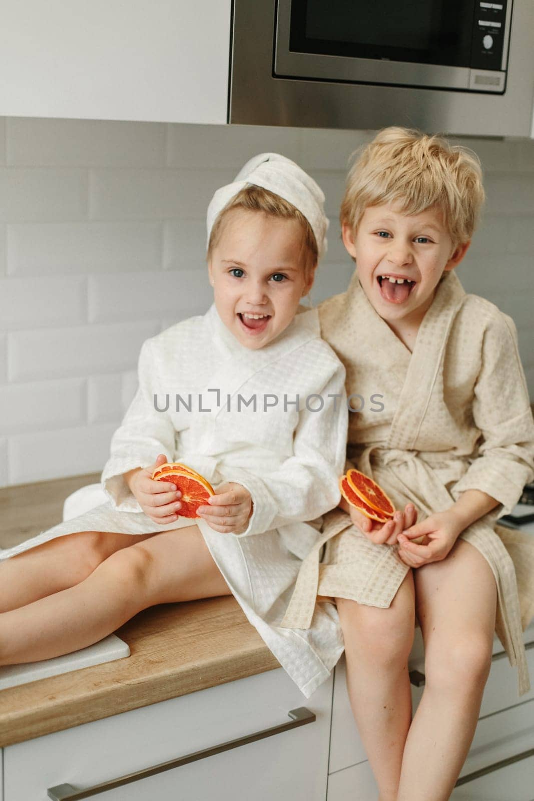 A girl and a boy in bathrobes are sitting in the kitchen with candied oranges in their hands, laughing.