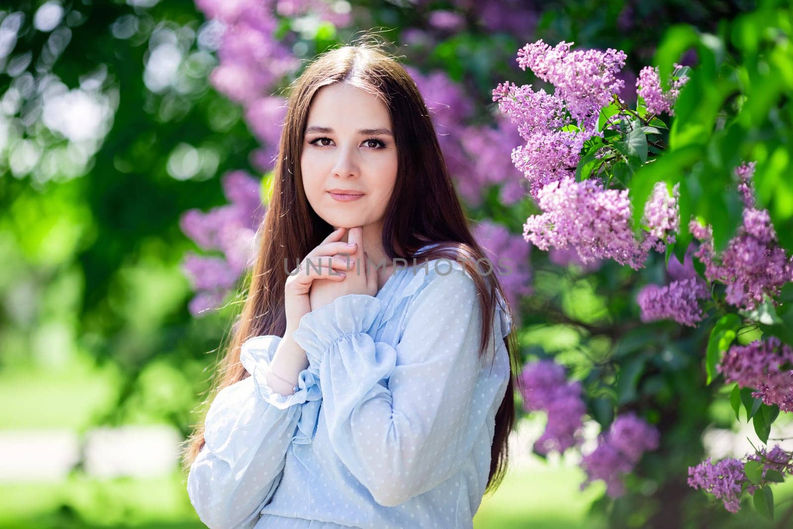 Adorable brunette girl with long hair stands with lilac flowers, in the garden, in sunny day, look at camera. Close up. copy space