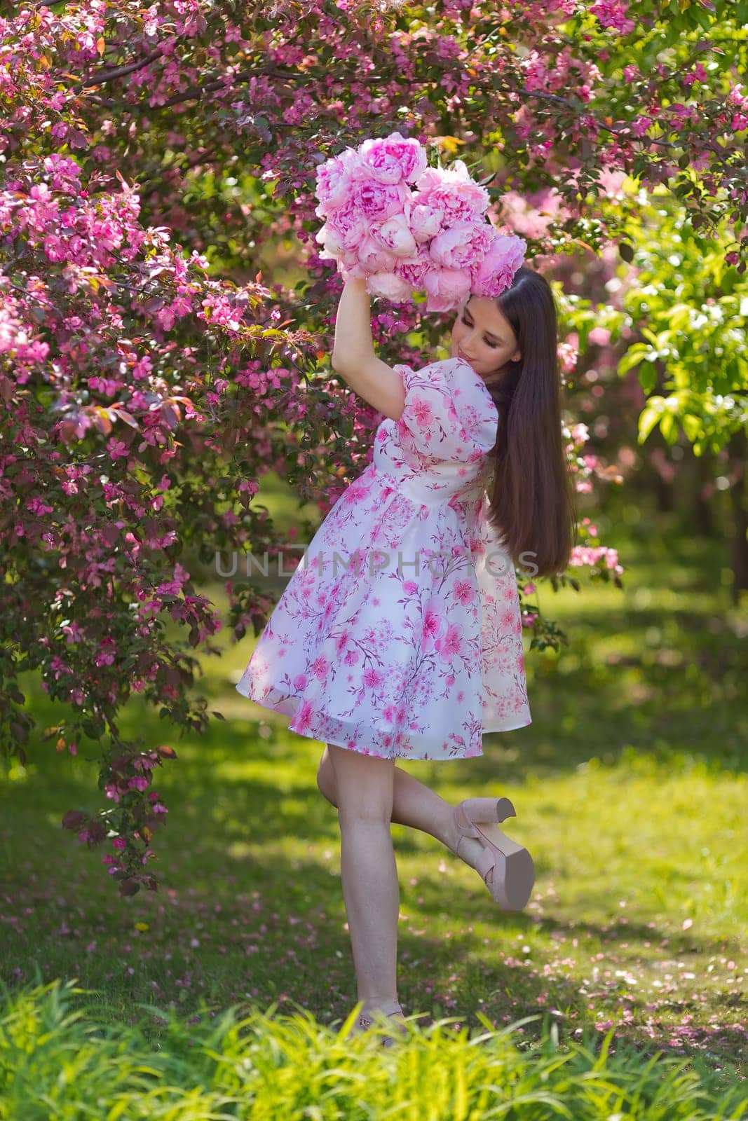 Happy beautiful girl with long hair, brunette, in a light pink dress, holding a bouquet of large pink peonies, standing near pink blossom in the garden on a sunny day. Space for copying. Vertical