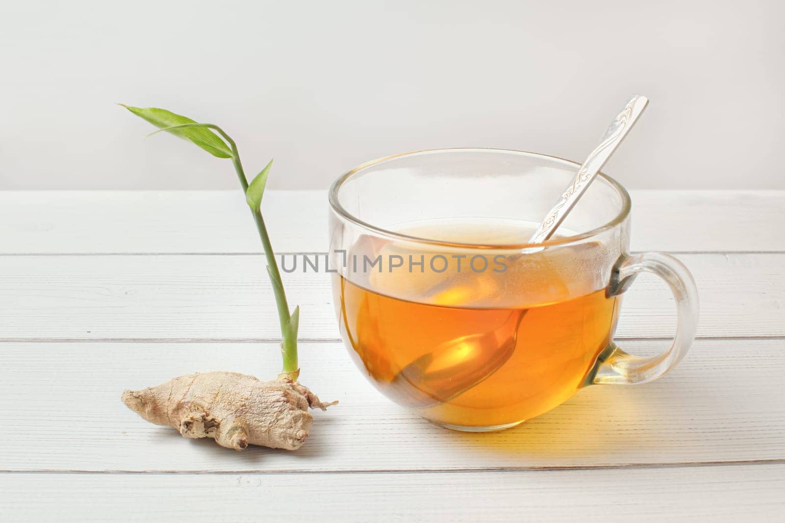 Spoon in glass cup of hot amber tea, dry ginger root with green sprout next to it.
