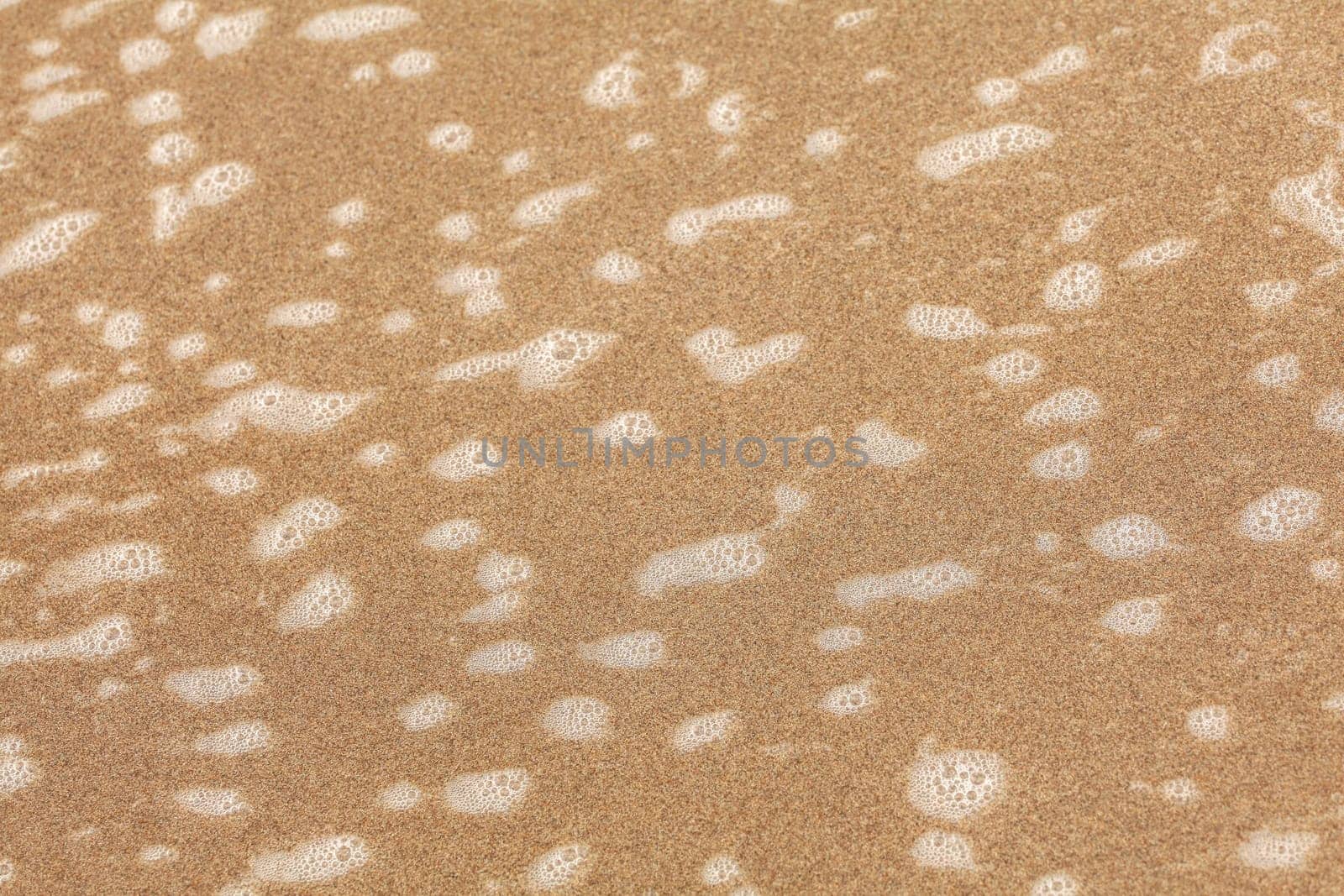 Small patches of white foam on beach sand wet from ocean water. Abstract sea / marine background.