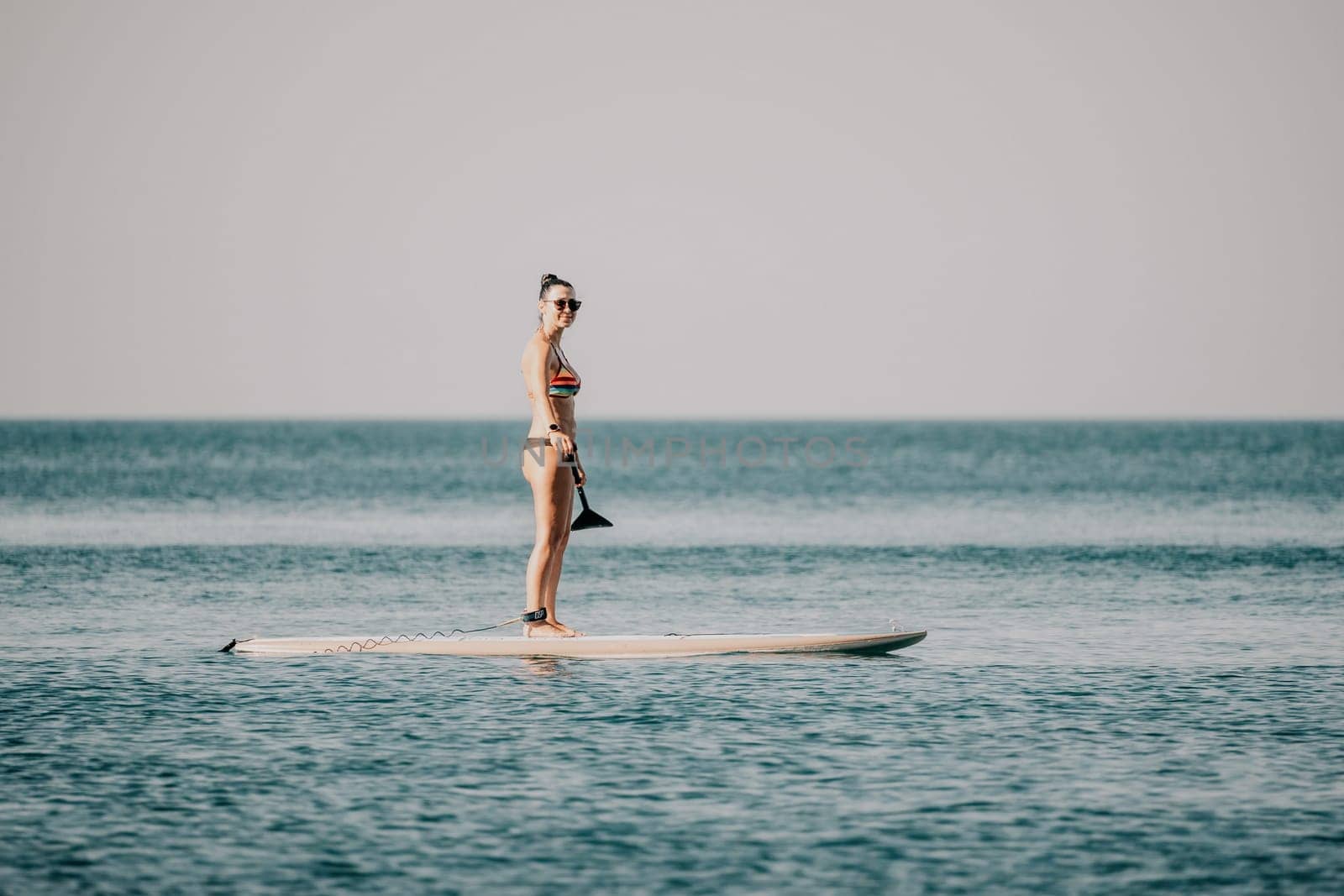 Sea woman sup. Silhouette of happy middle aged tanned woman in rainbow bikini, surfing on SUP board, confident paddling through water surface. Idyllic sunset. Active lifestyle at sea or river. by panophotograph