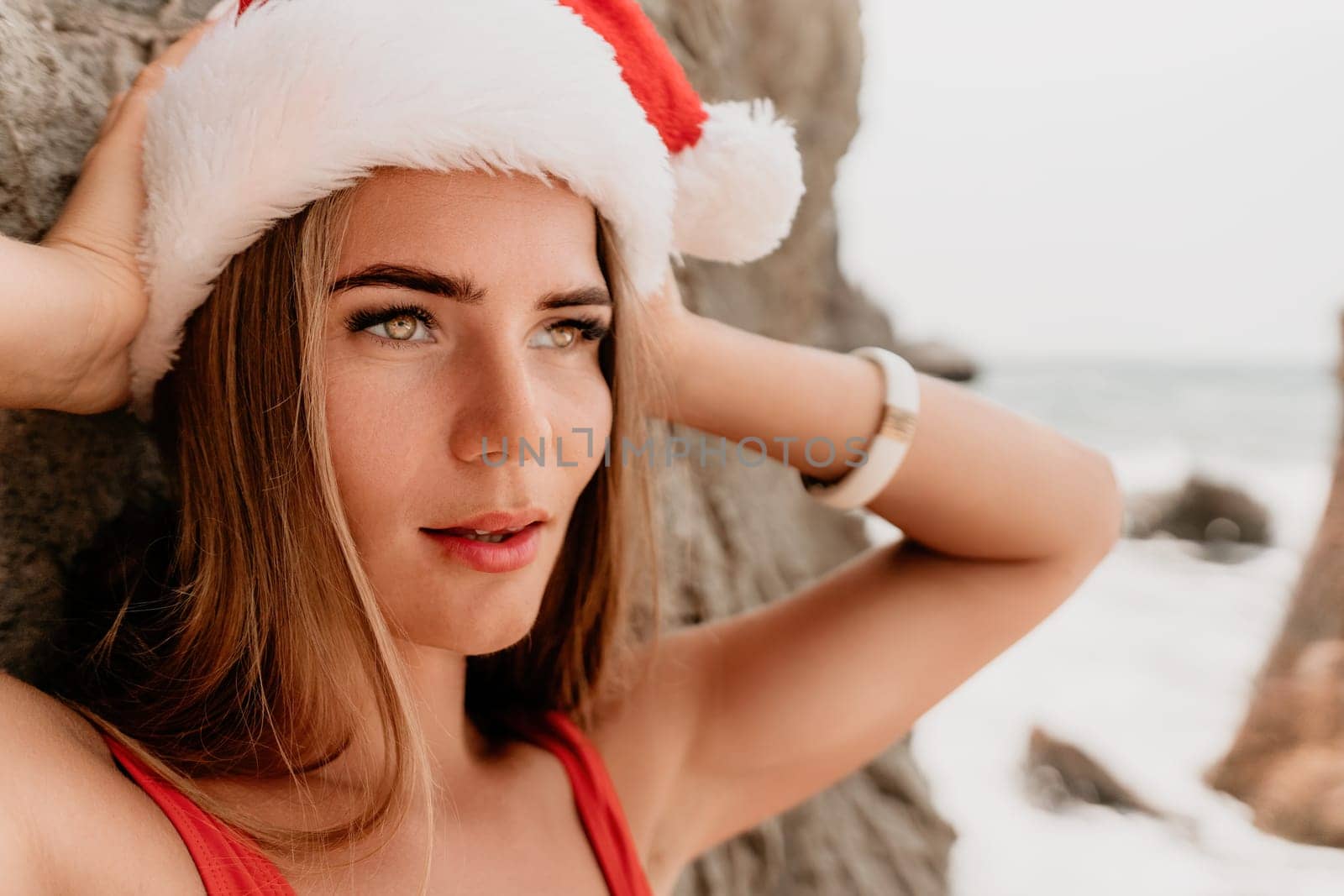 Woman summer travel sea. Happy tourist in red bikini and Santas hat enjoy taking picture outdoors for memories. Woman traveler posing on the beach surrounded by volcanic mountains, sharing travel joy by panophotograph