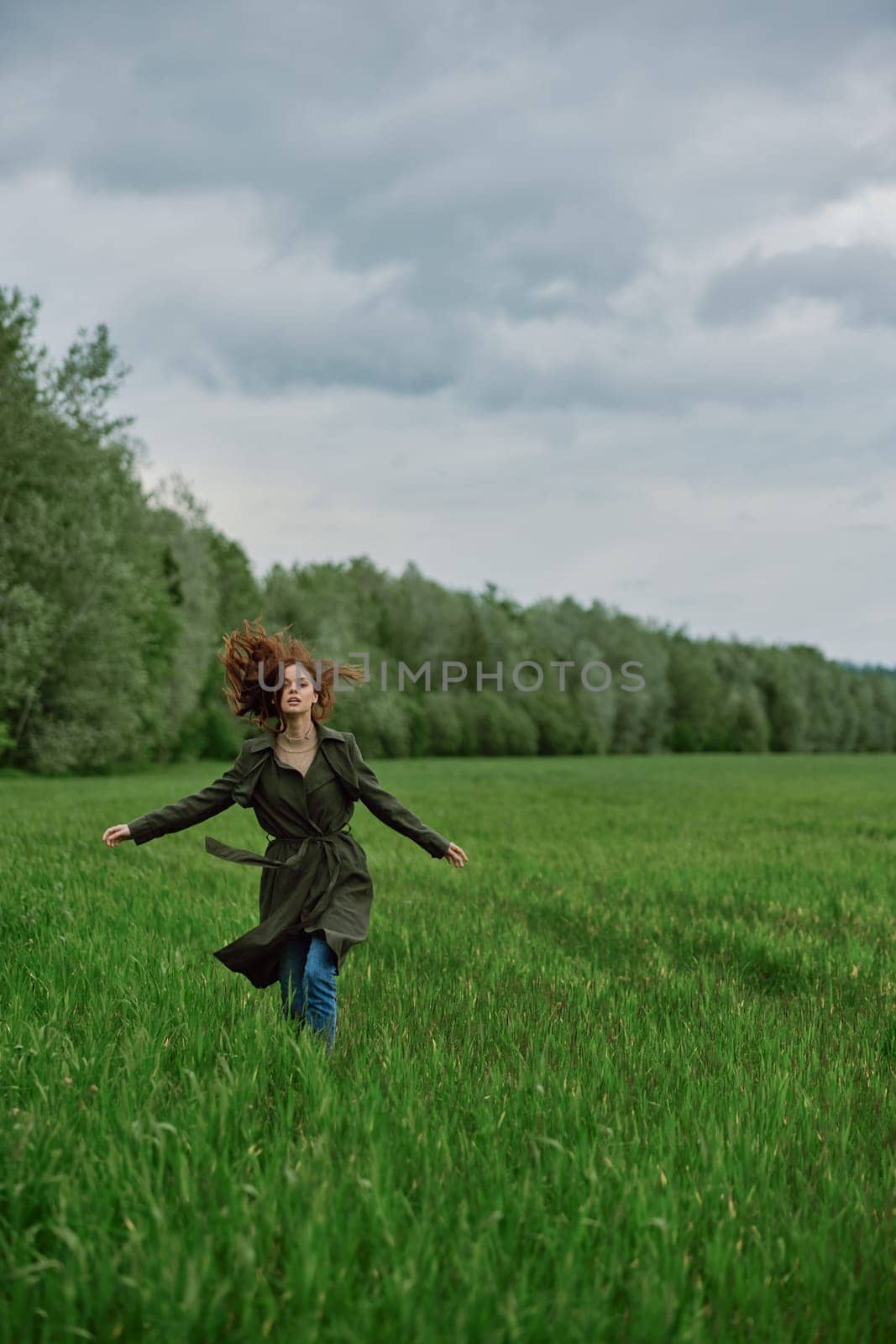 a beautiful woman in a long raincoat runs across a field in high grass in spring in cloudy weather. High quality photo