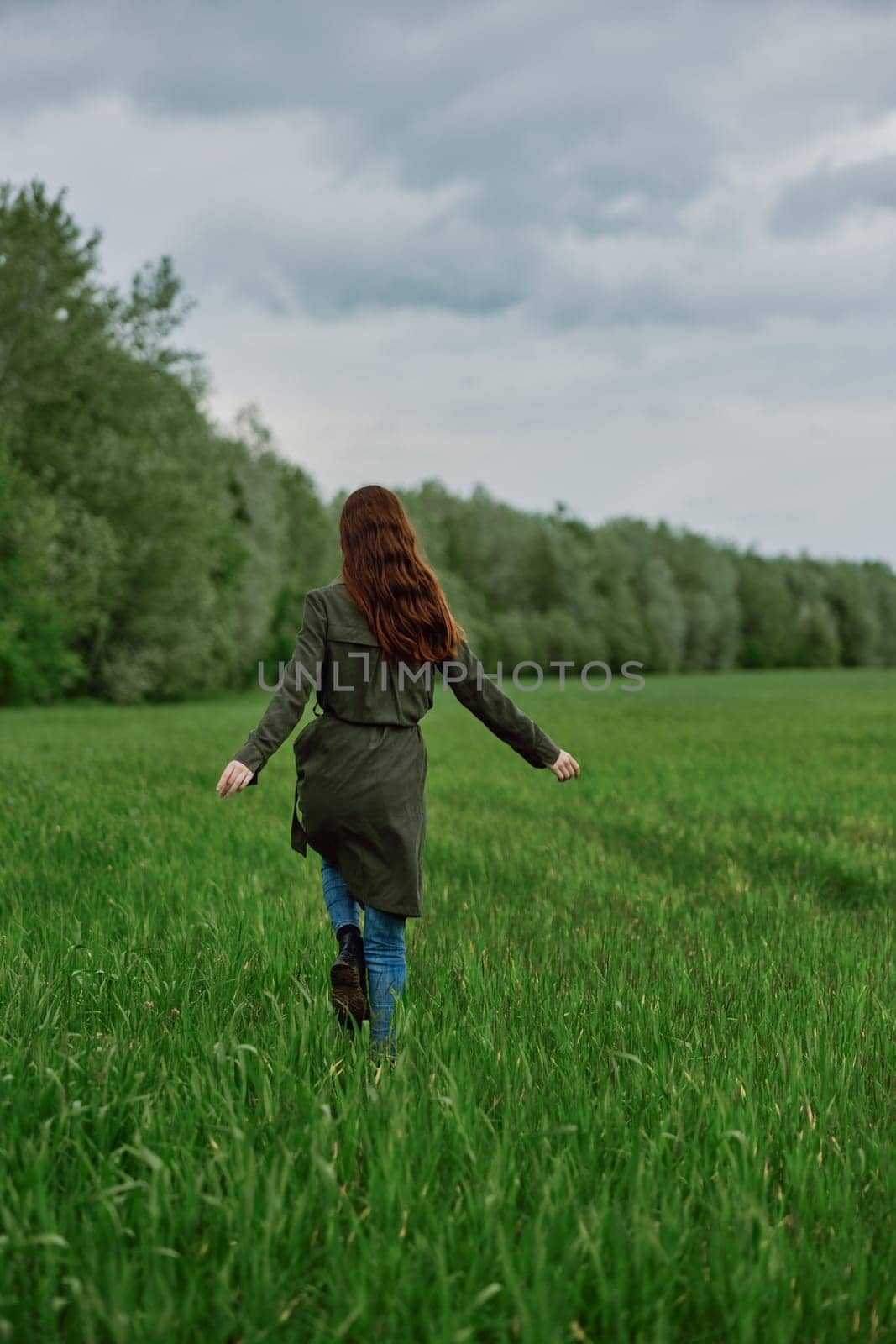 a woman in a long raincoat runs across a field in tall green grass in cloudy weather in spring. High quality photo