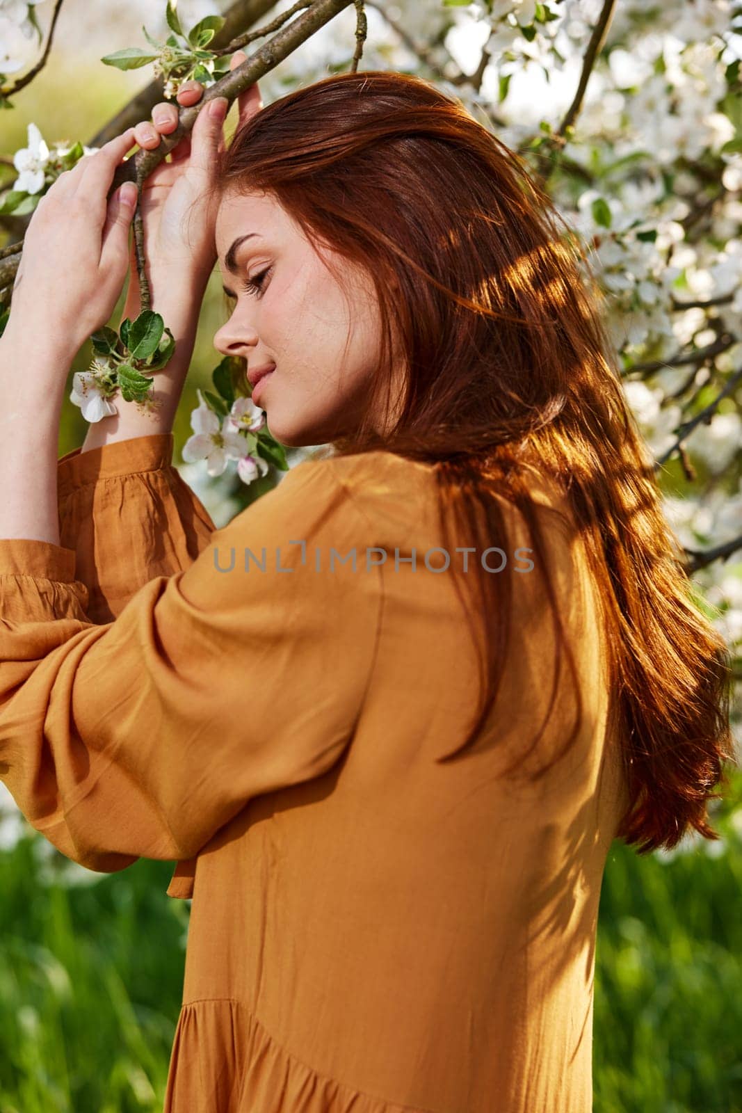 a happy, modest woman is standing in an orange dress near a flowering tree and holding her hands on the branches leans her head to them by Vichizh