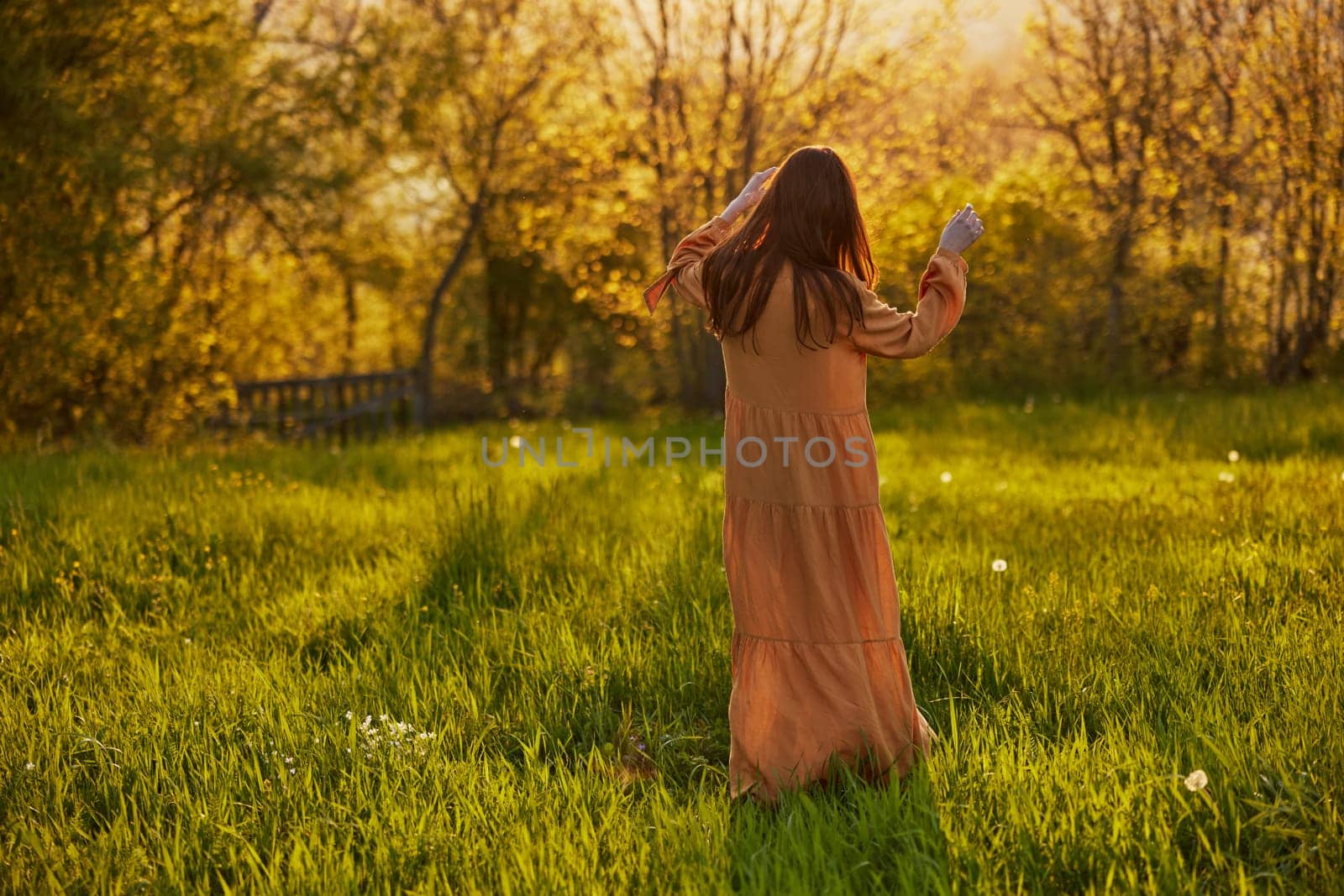 a slender woman with long hair stands in a field with her back to the camera, illuminated by the rays of the setting sun and happily poses enjoying the warm weather and rest by Vichizh