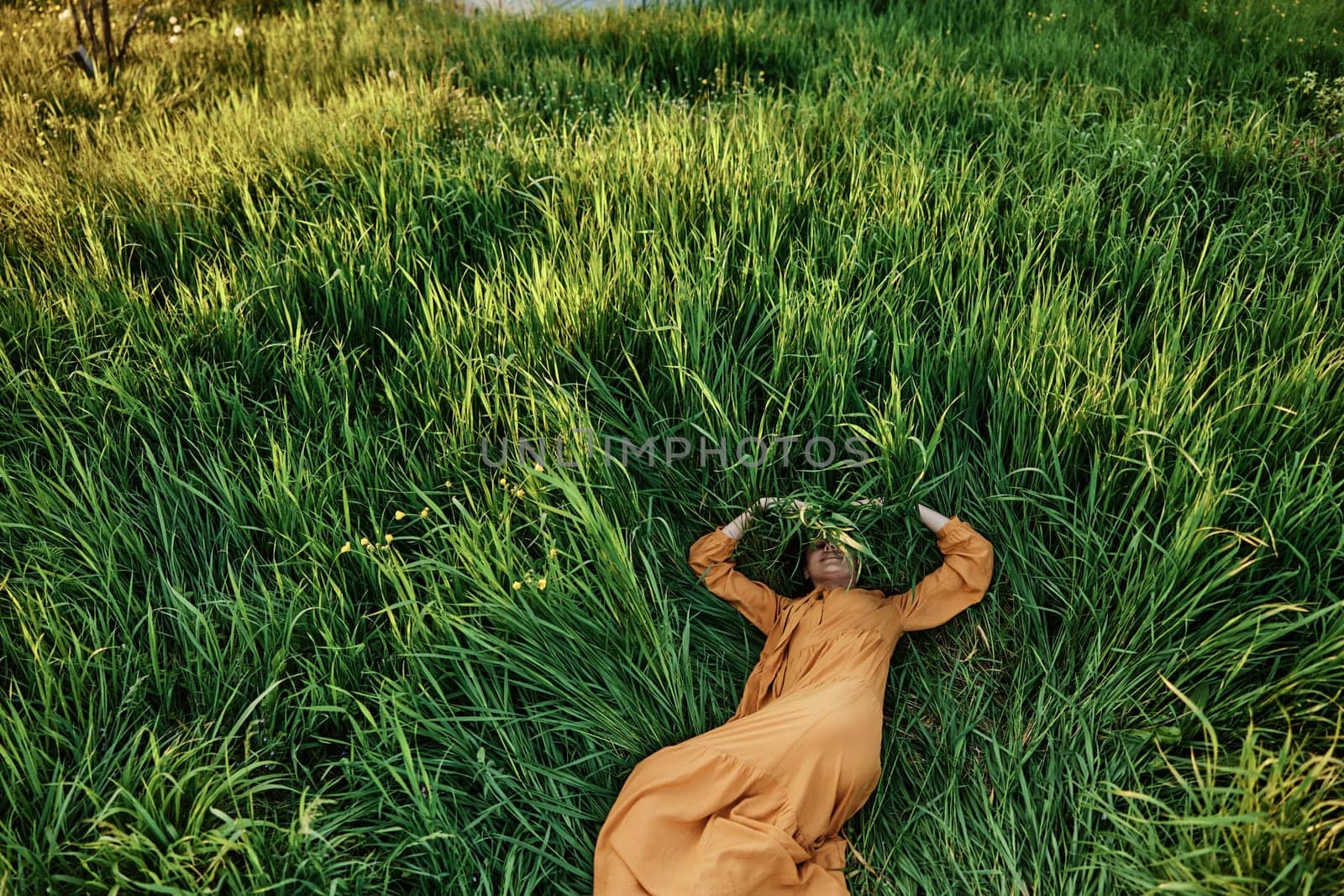 a sweet, calm woman in an orange dress lies in a green field, covering her face with long leaves of grass, enjoying silence and peace. Horizontal photo taken from above by Vichizh