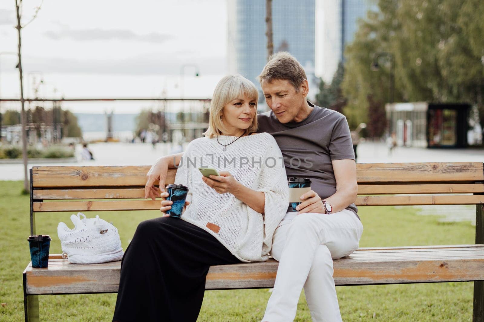 An adult mature happy couple in love sitting on bench with phone outdoors in park. A blonde caucasian man and woman spend time together and drinking coffee. Senior wife and husband walking outside