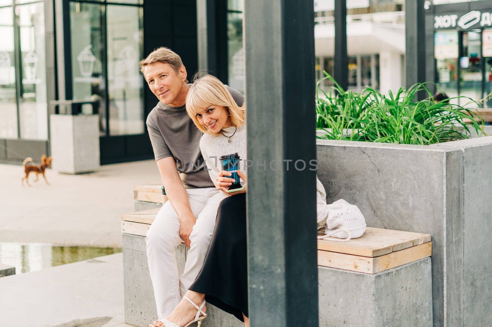 An adult mature happy couple in love sitting on bench outdoors in city street park. A blonde caucasian man and woman spend time together and drinking coffee. Senior wife and husband walking outside. by Ostanina