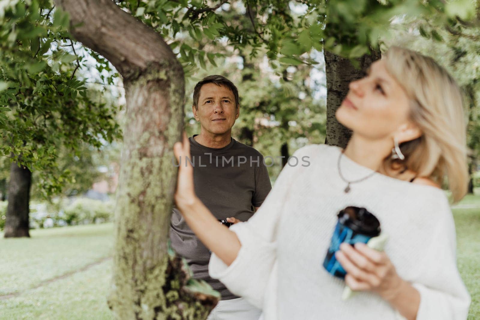An adult mature happy couple in love hugging outdoors in city park. A blonde caucasian man and woman spend time together and drinking coffee. Senior wife and husband walking outside. by Ostanina