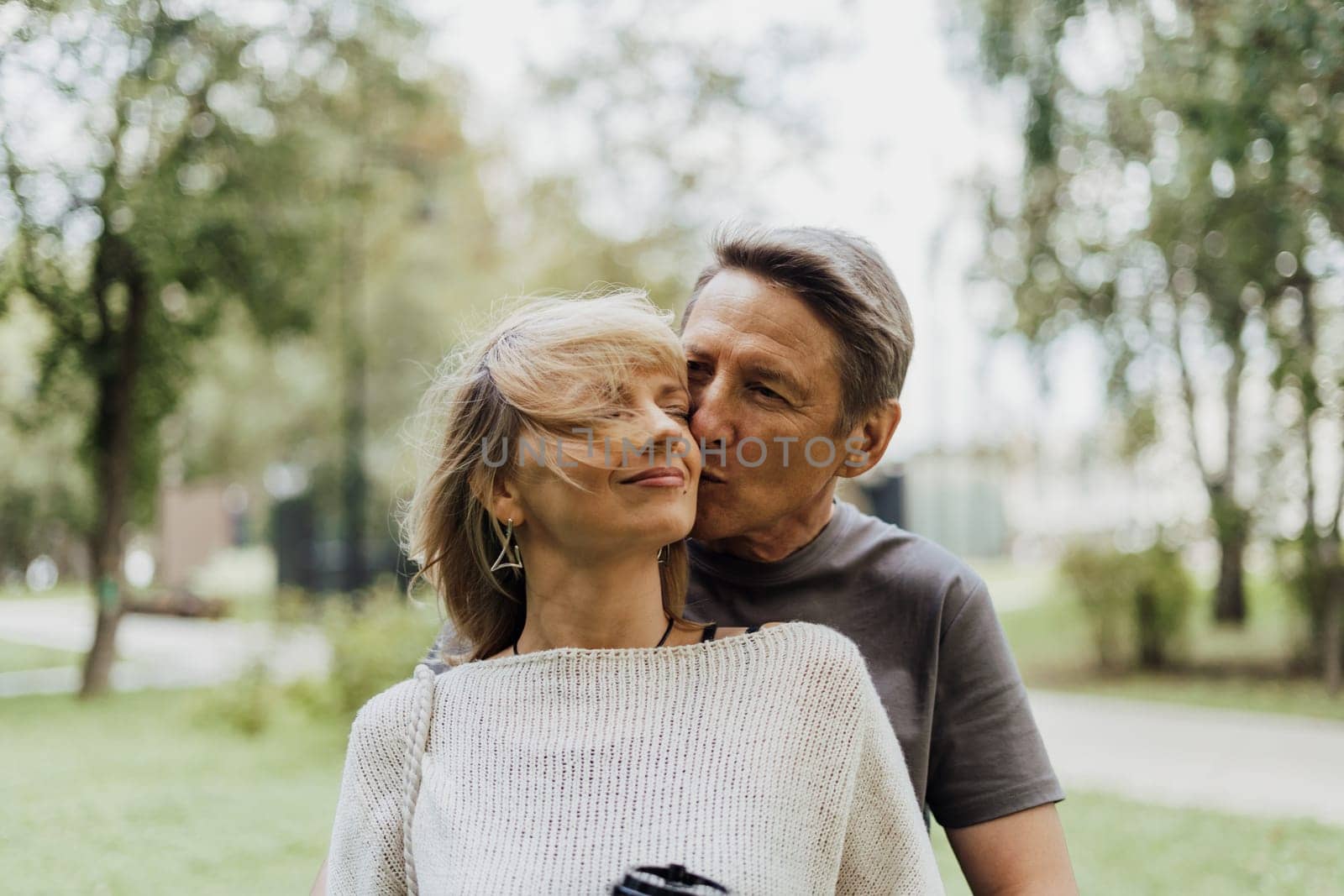 An adult mature happy couple in love hugging outdoors in city park. A blonde caucasian man and woman spend time together and drinking coffee. Senior wife and husband walking outside. by Ostanina