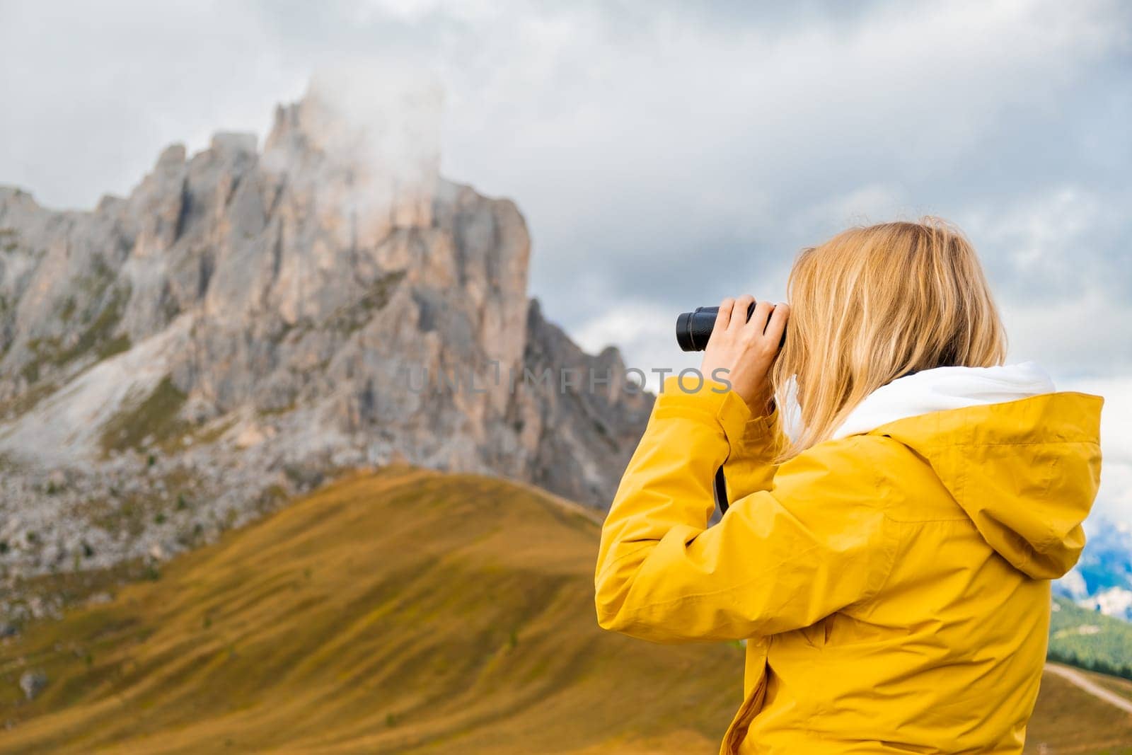 Young blonde woman looks in binoculars to high mountain pass in the Dolomites, the Giau Pass.