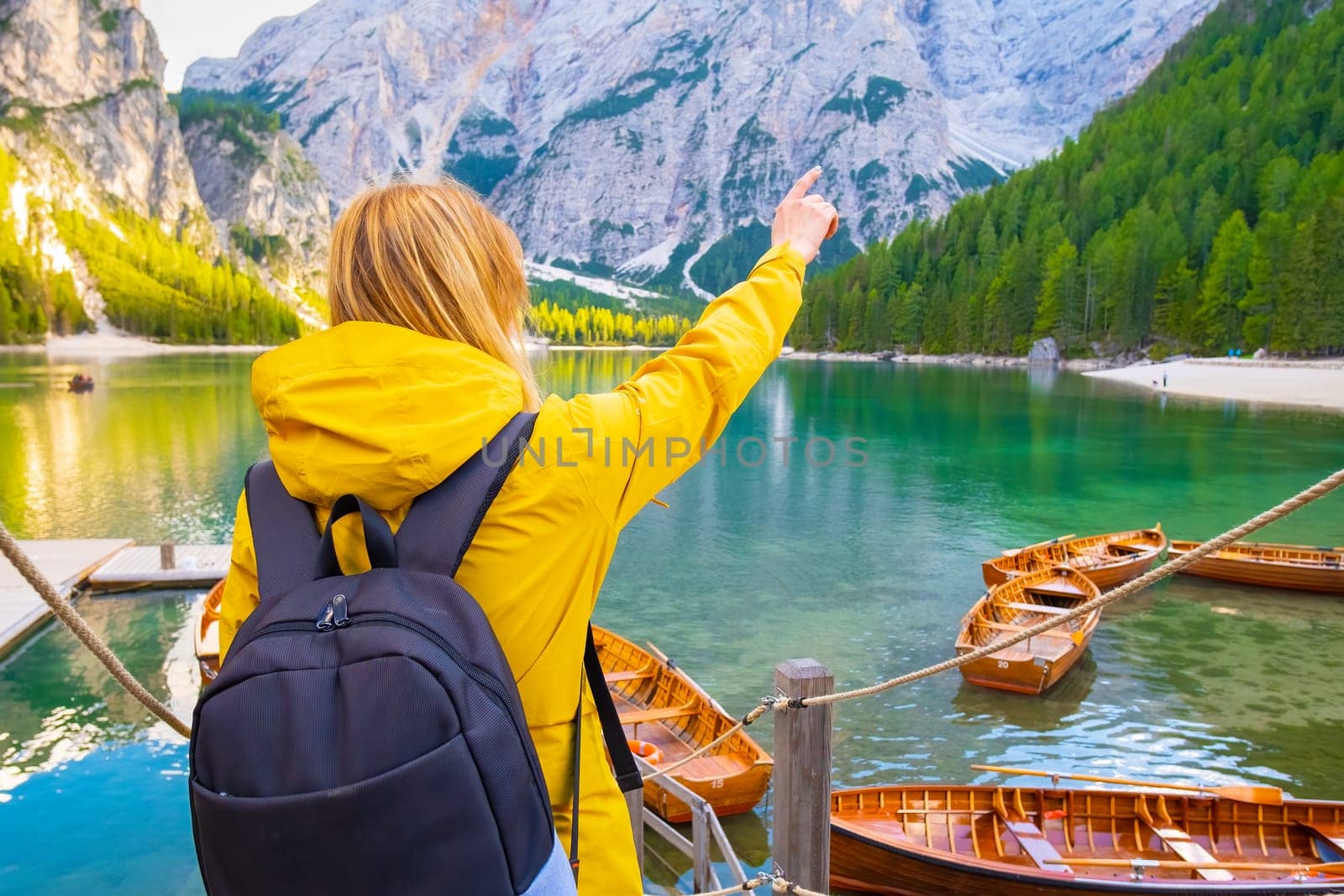 Tourist wearing yellow sport jacket shows on the mountain by finger on the lake Braies with wooden boats. Woman with backpack enjoying landscape of Dolomites