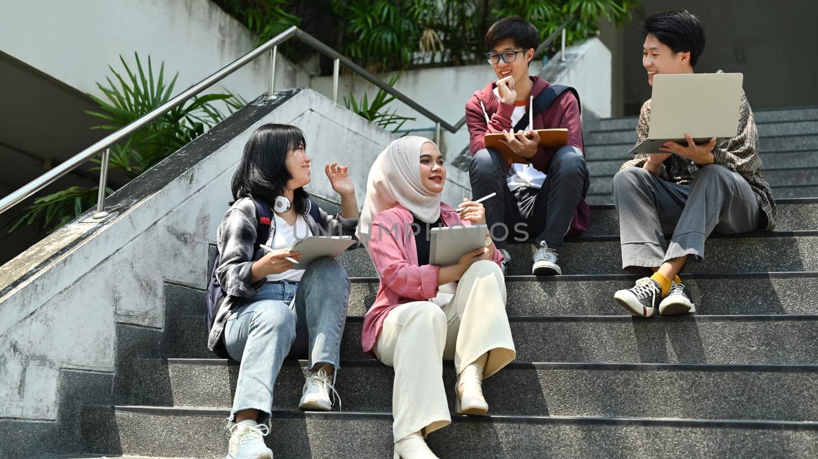 Happy diverse group of college students sitting on stairs working together on study project. Education and youth lifestyle concept.