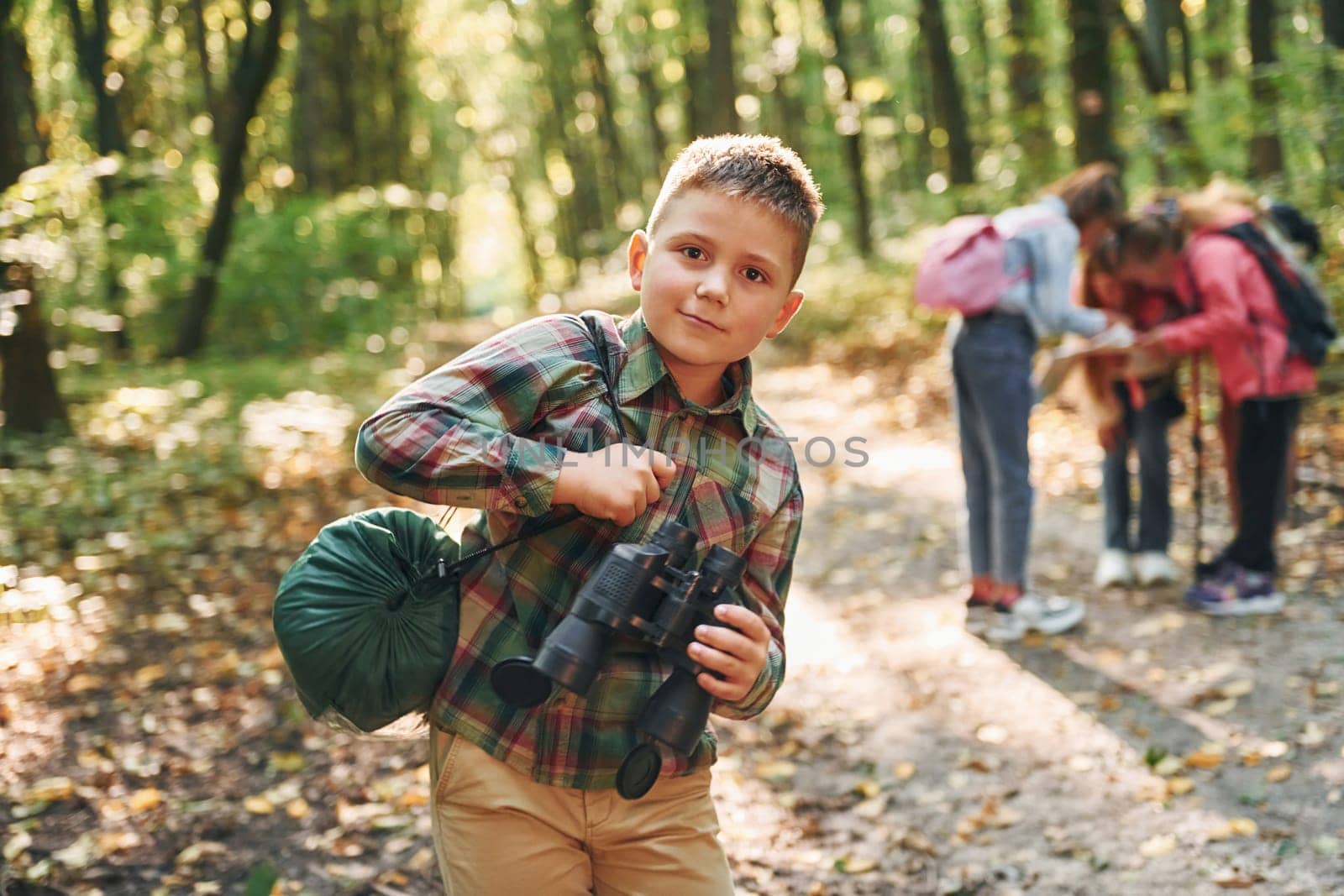 Boy with binoculars standing in front of his friends. Kids in green forest at summer daytime together by Standret