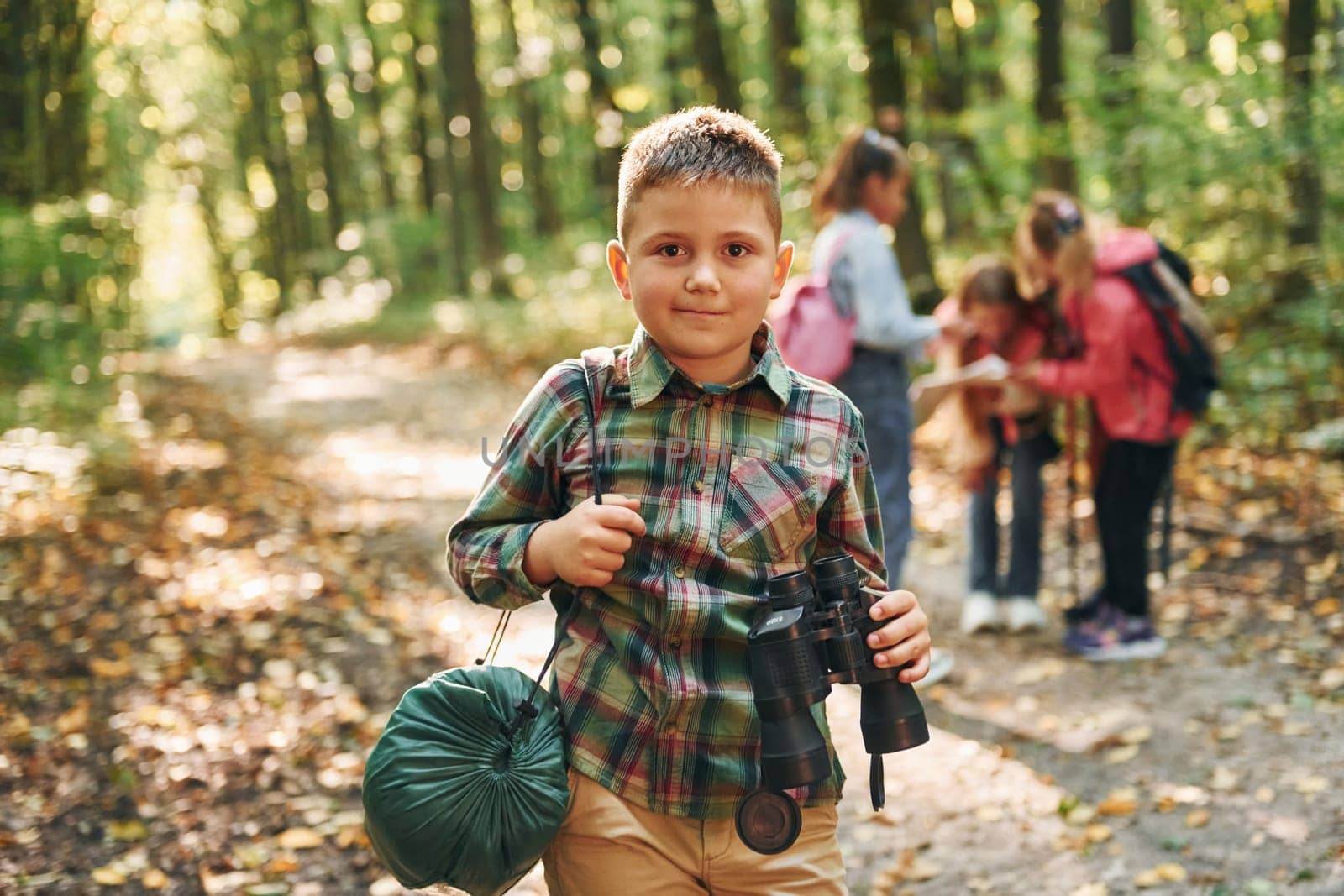 Boy with binoculars standing in front of his friends. Kids in green forest at summer daytime together by Standret