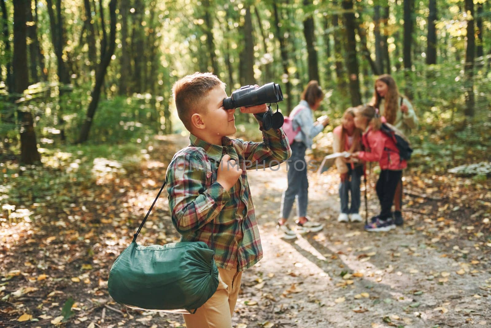 Boy with binoculars standing in front of his friends. Kids in green forest at summer daytime together by Standret