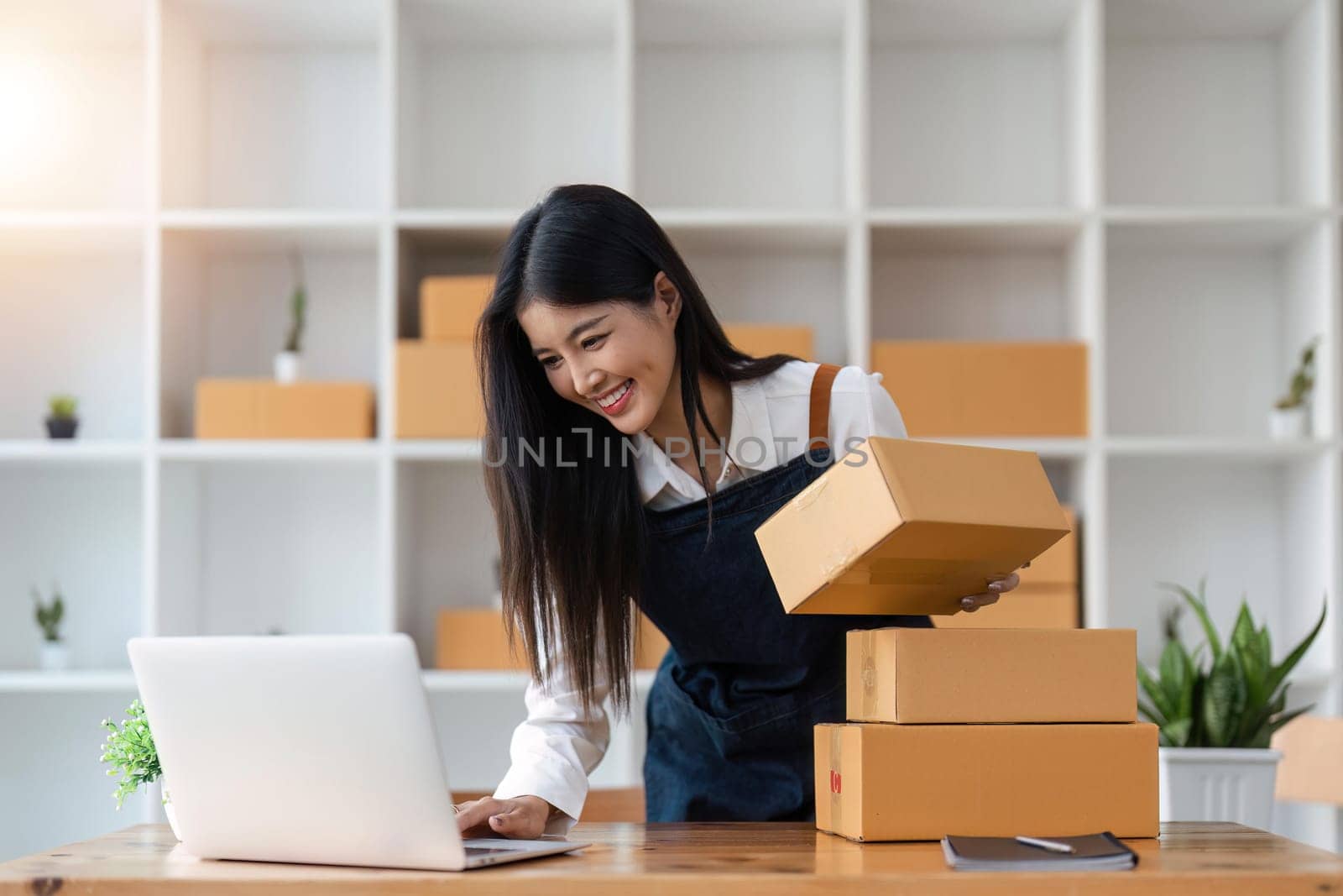 Startup SME small business entrepreneur of freelance Asian woman using laptop and box to receive and review orders online to prepare to pack sell to customers, online sme business ideas by nateemee