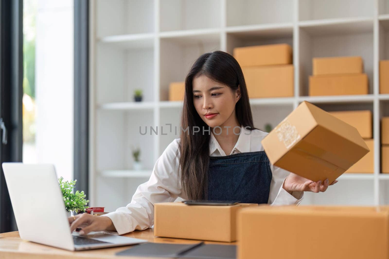 Startup SME small business entrepreneur of freelance Asian woman using laptop and box to receive and review orders online to prepare to pack sell to customers, online sme business ideas.