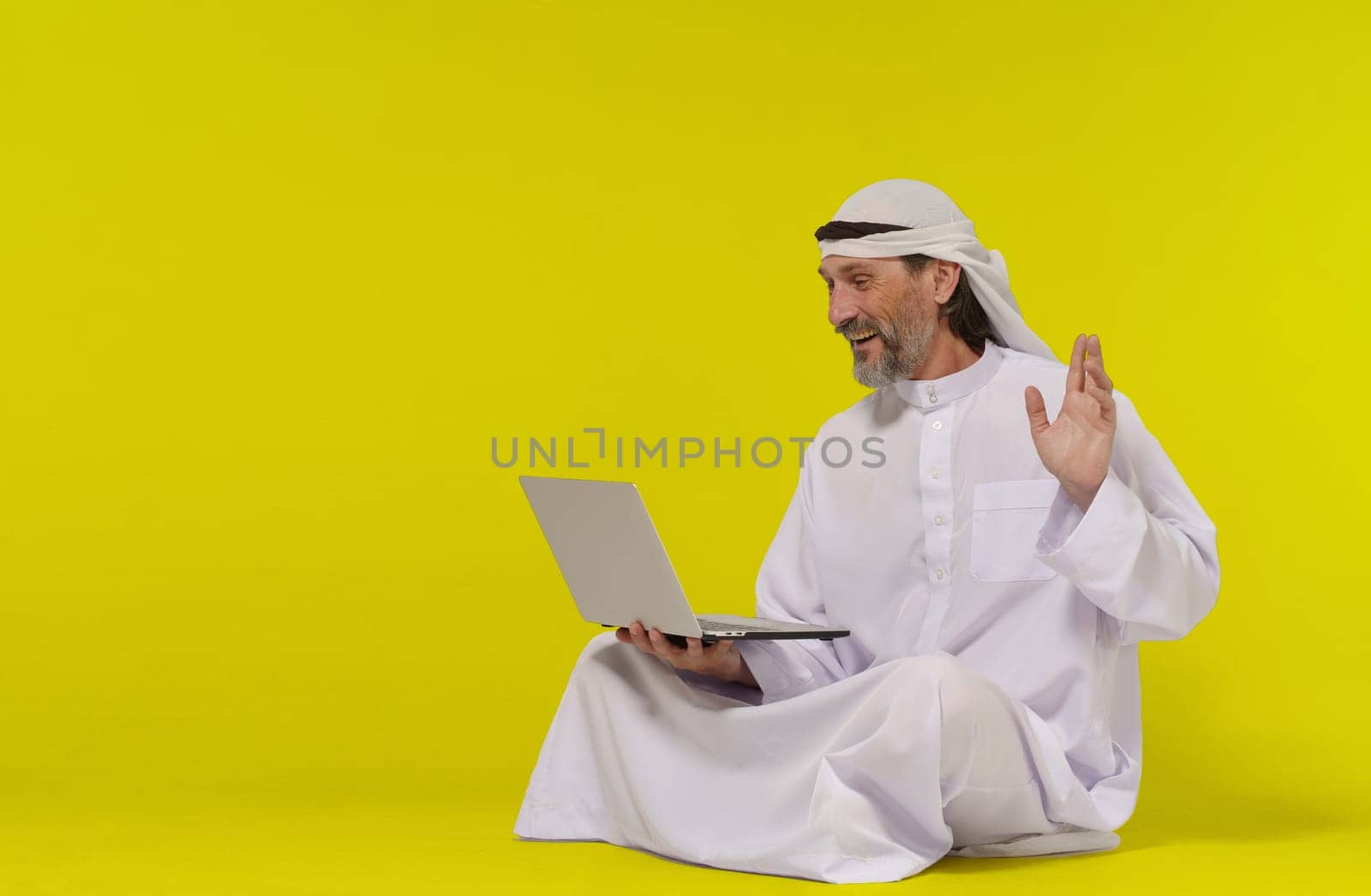Concept online communication. Muslim person sitting on floor greets someone with their palm raised while holding a laptop in their hand. Copy space allows for easy customization and versatility and global connection through technology. High quality photo