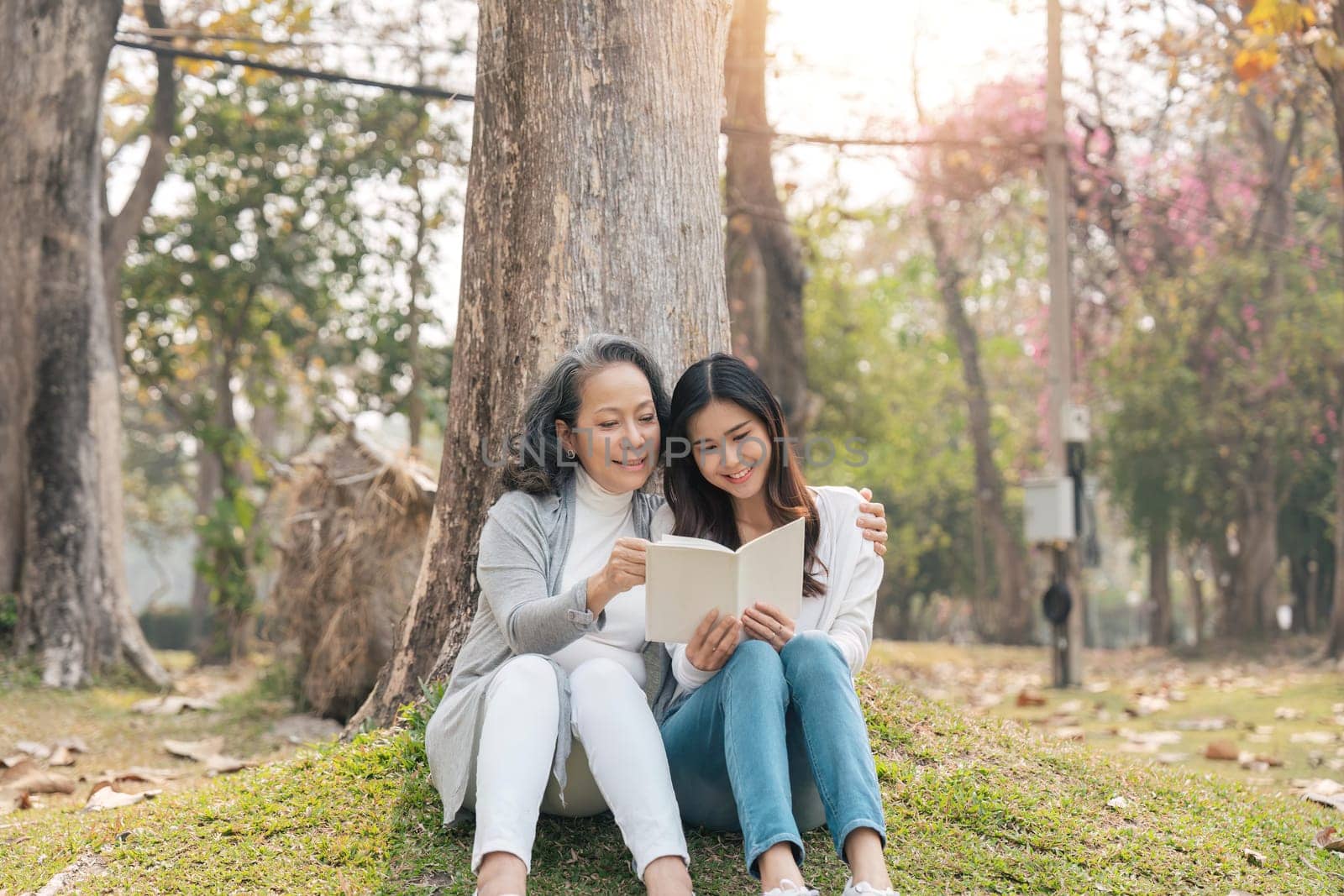 Lovely granddaughter and grandmother sitting under the tree in the green park, read a book together.