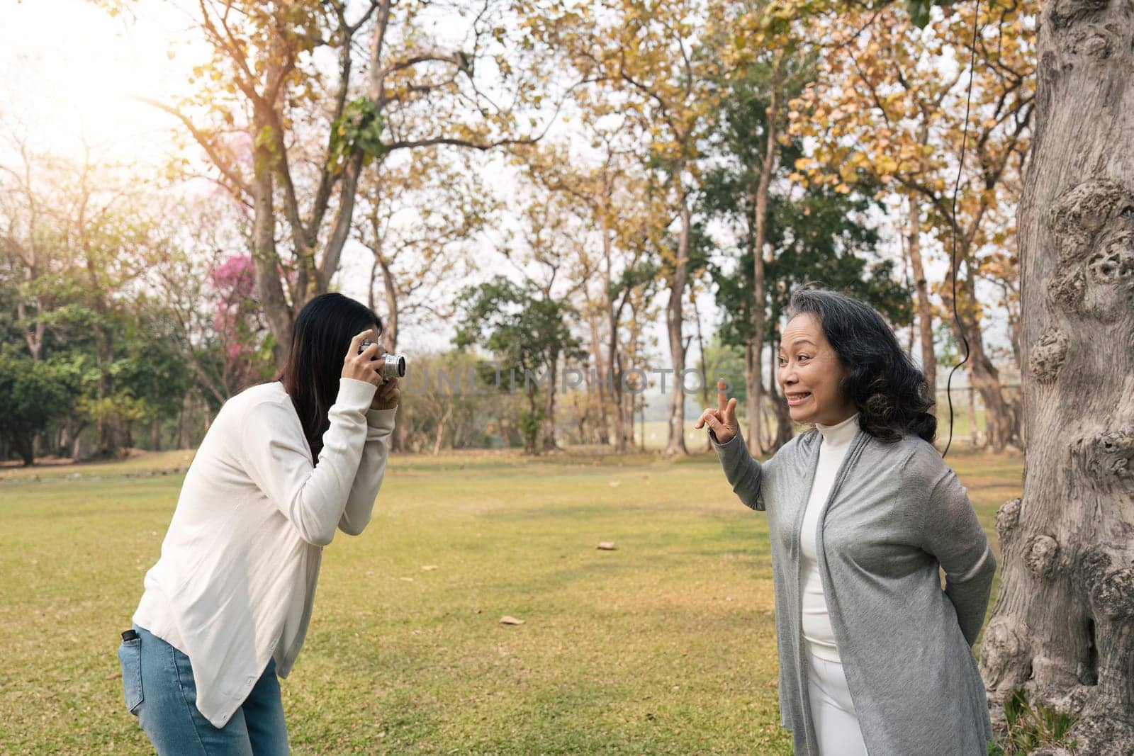 Lovely Asian granddaughter taking a picture of her happy grandmother with camera while strolling in the green park together.