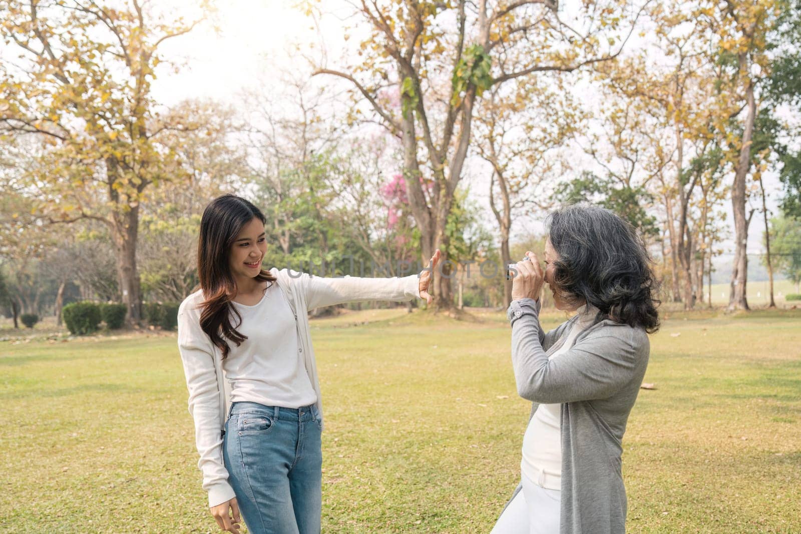 Happy grandmother taking a picture of her lovely granddaughter with a camera while strolling in the green park together.