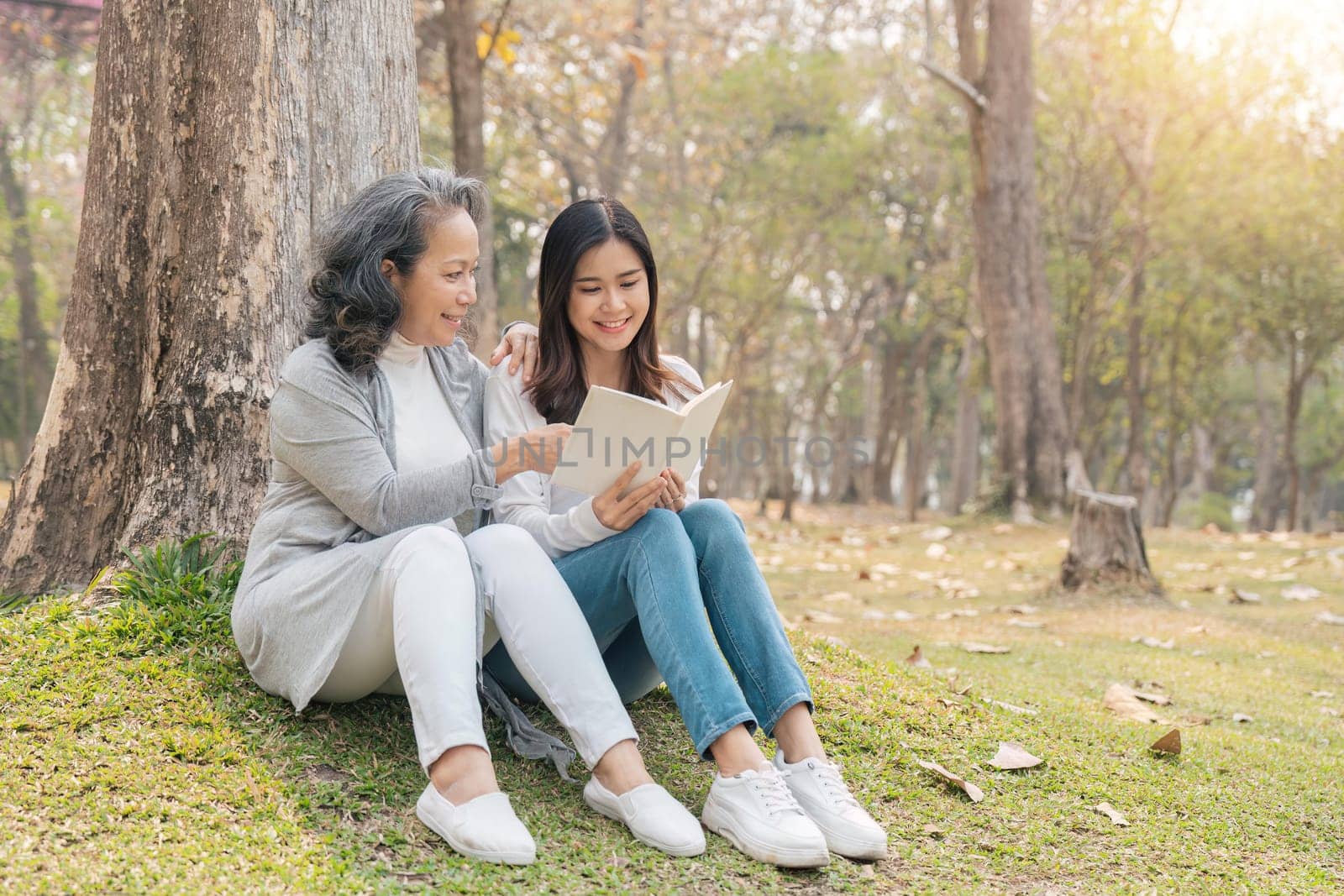 Lovely granddaughter and grandmother sitting under the tree in the green park, read a book together.