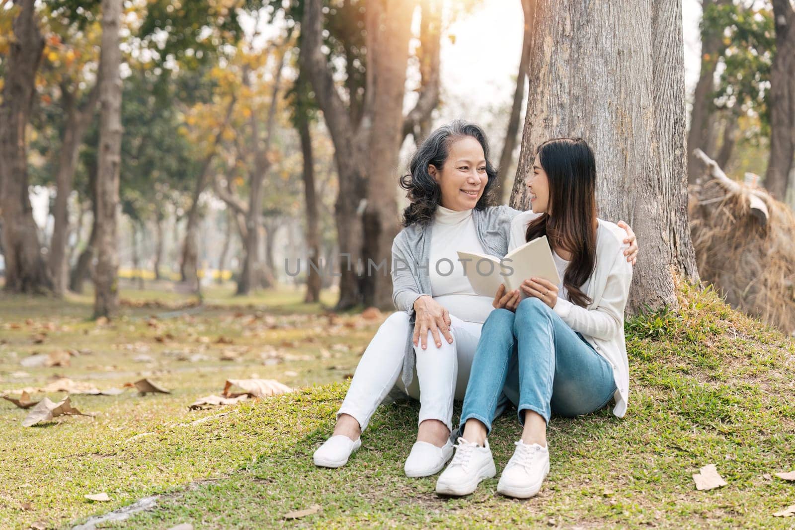 Lovely granddaughter and grandmother sitting under the tree in the green park, read a book together.